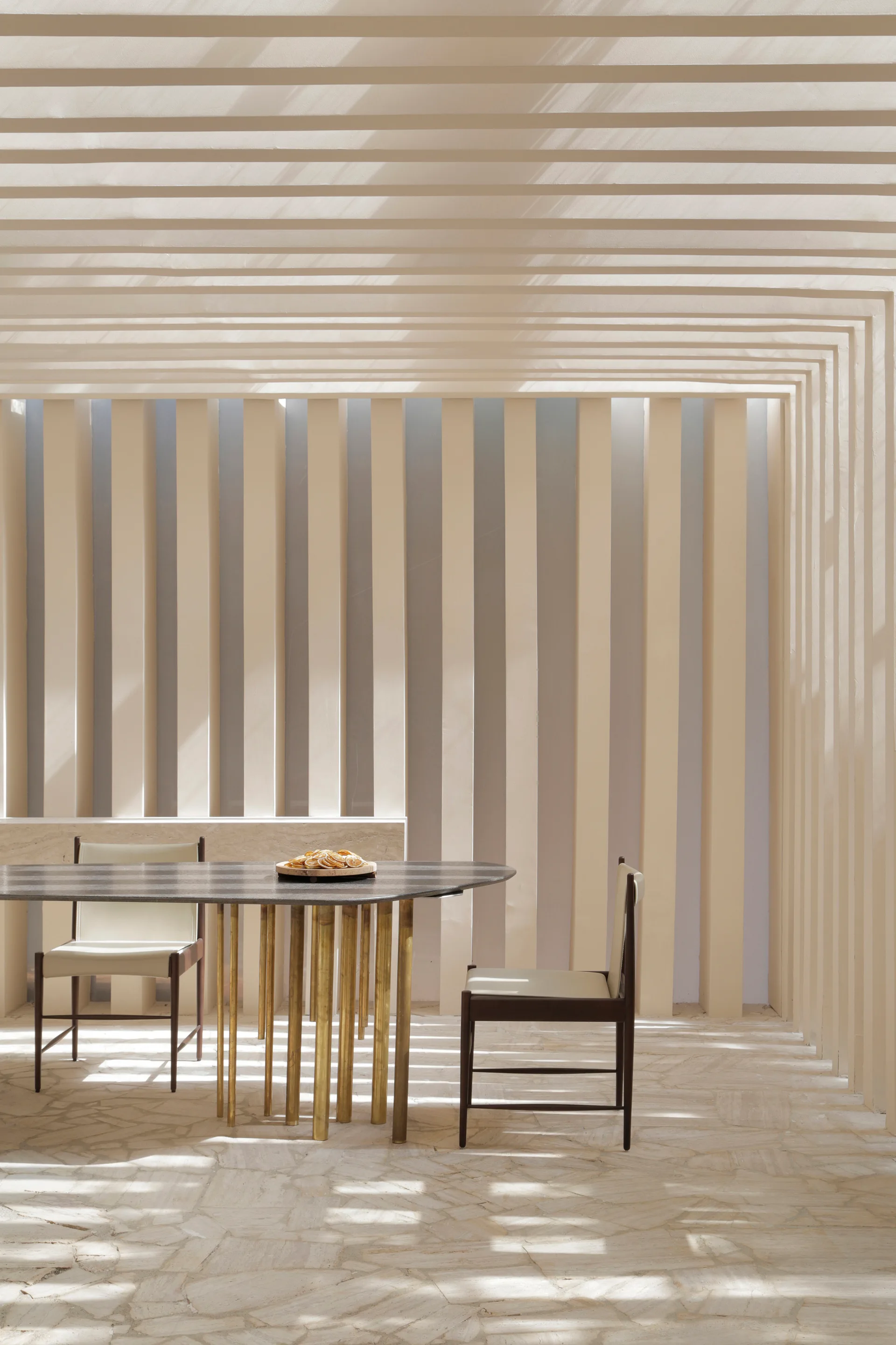 An outdoor patio dining table with metallic legs stands on a stone floor under a continuous beige slatted wall and pergola ceiling.