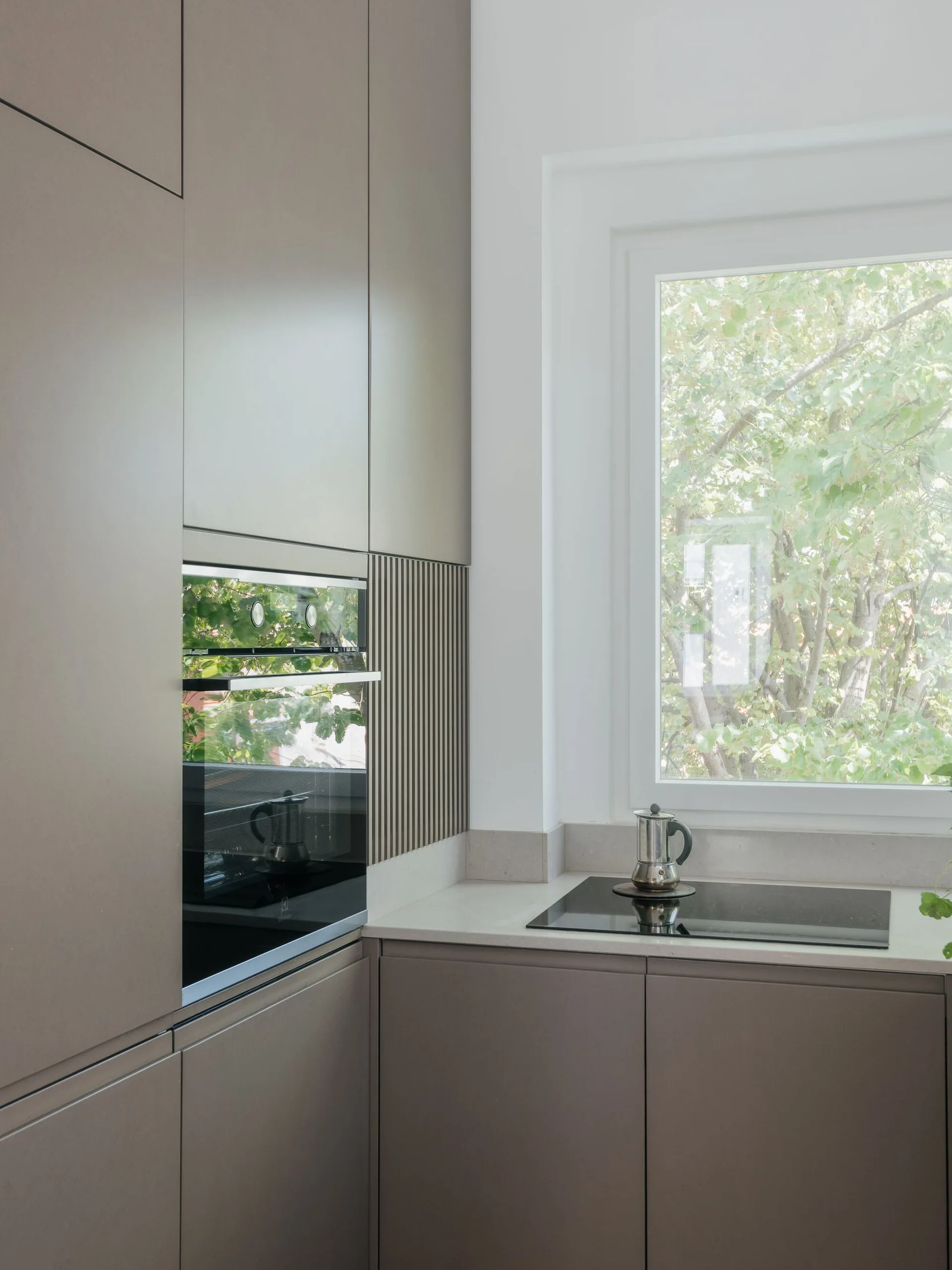 Matte taupe kitchen cabinets with an integrated oven and a stone countertop sit next to a white-framed window looking out at leafy green trees.