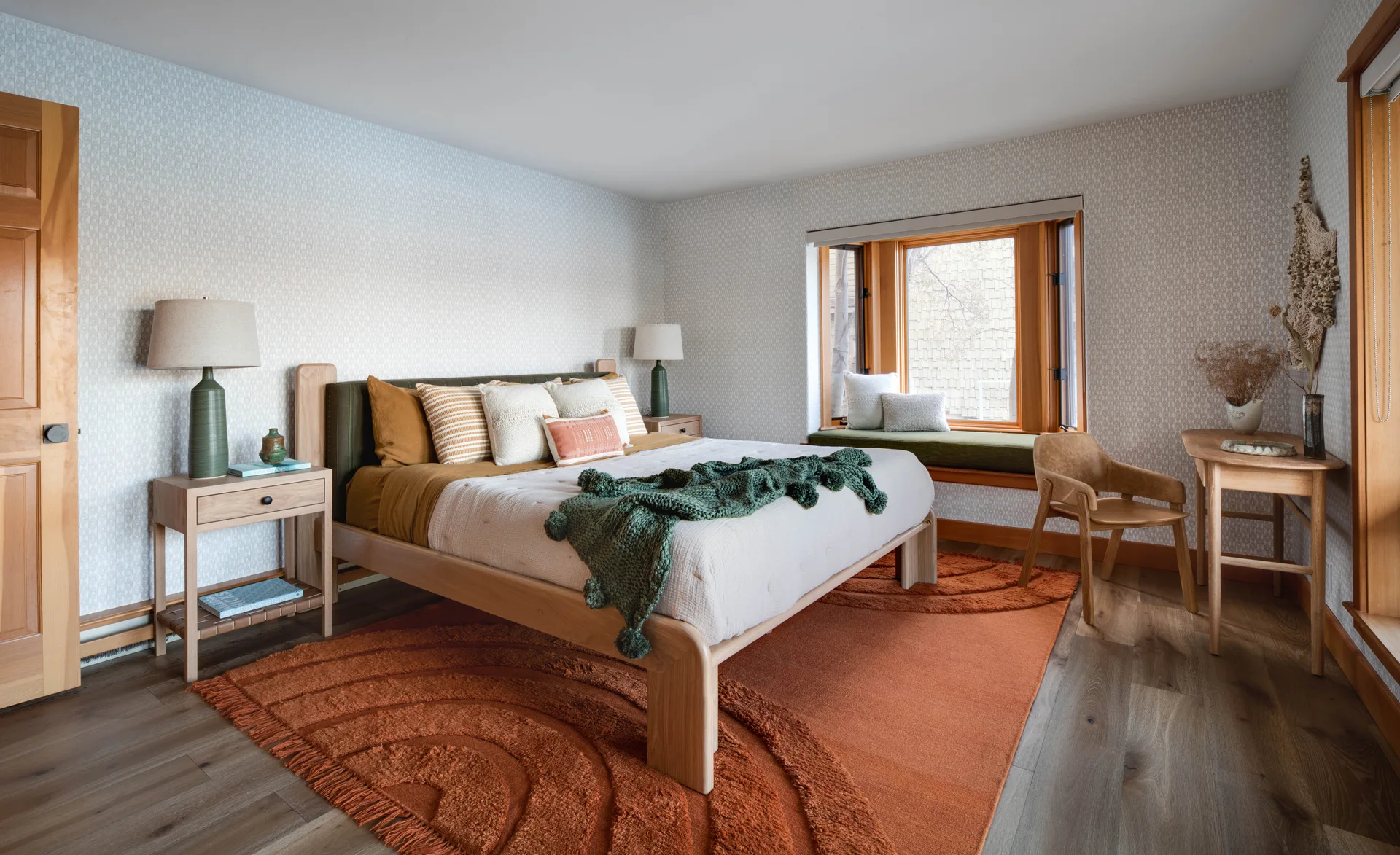 A bedroom features a wooden bed with earthy linens, a large terracotta rug, and a bay window seat set against patterned wallpaper.