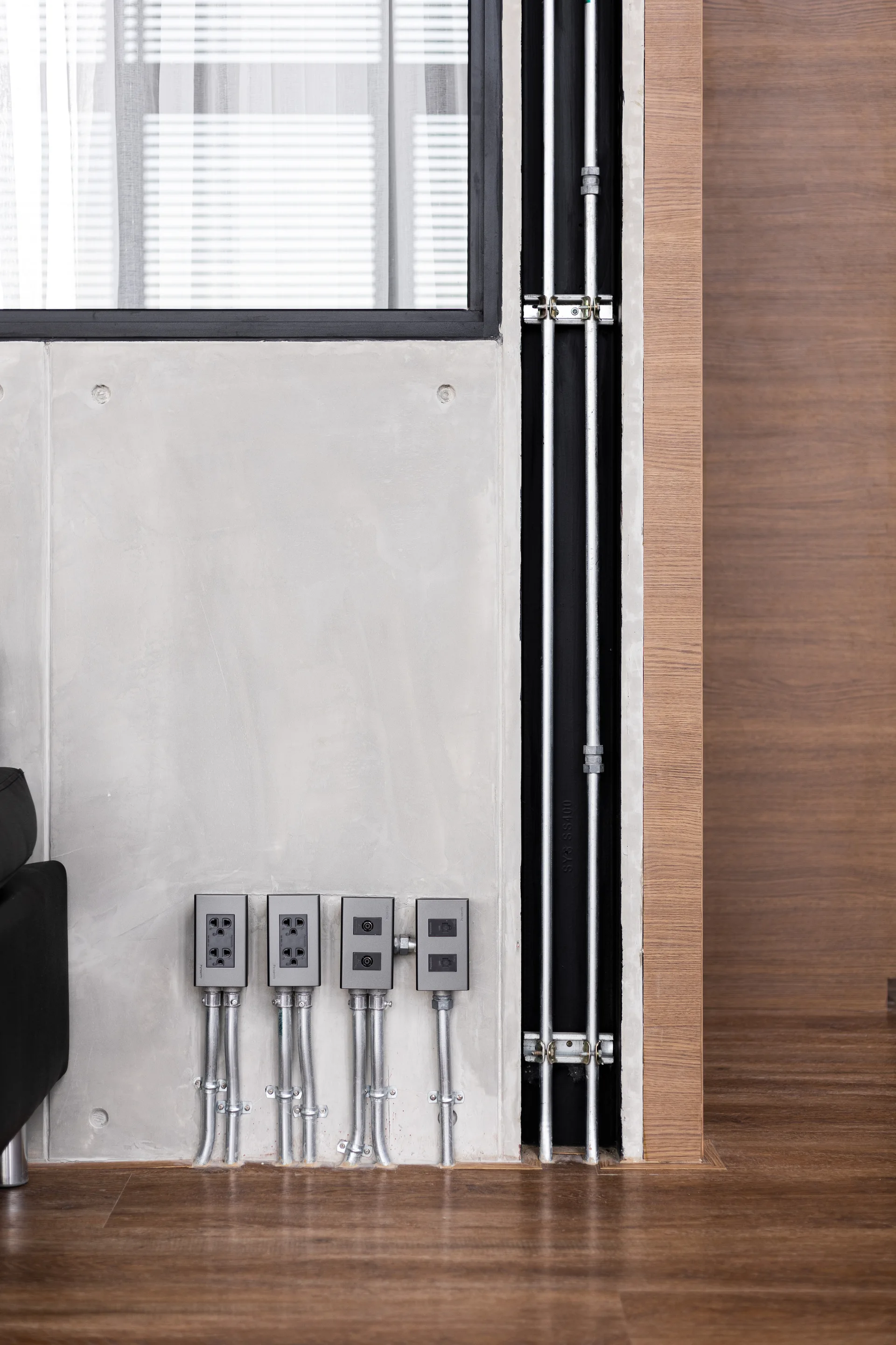 A concrete interior wall features four exposed silver electrical outlets and vertical metal conduits next to a wood panel and wood flooring.