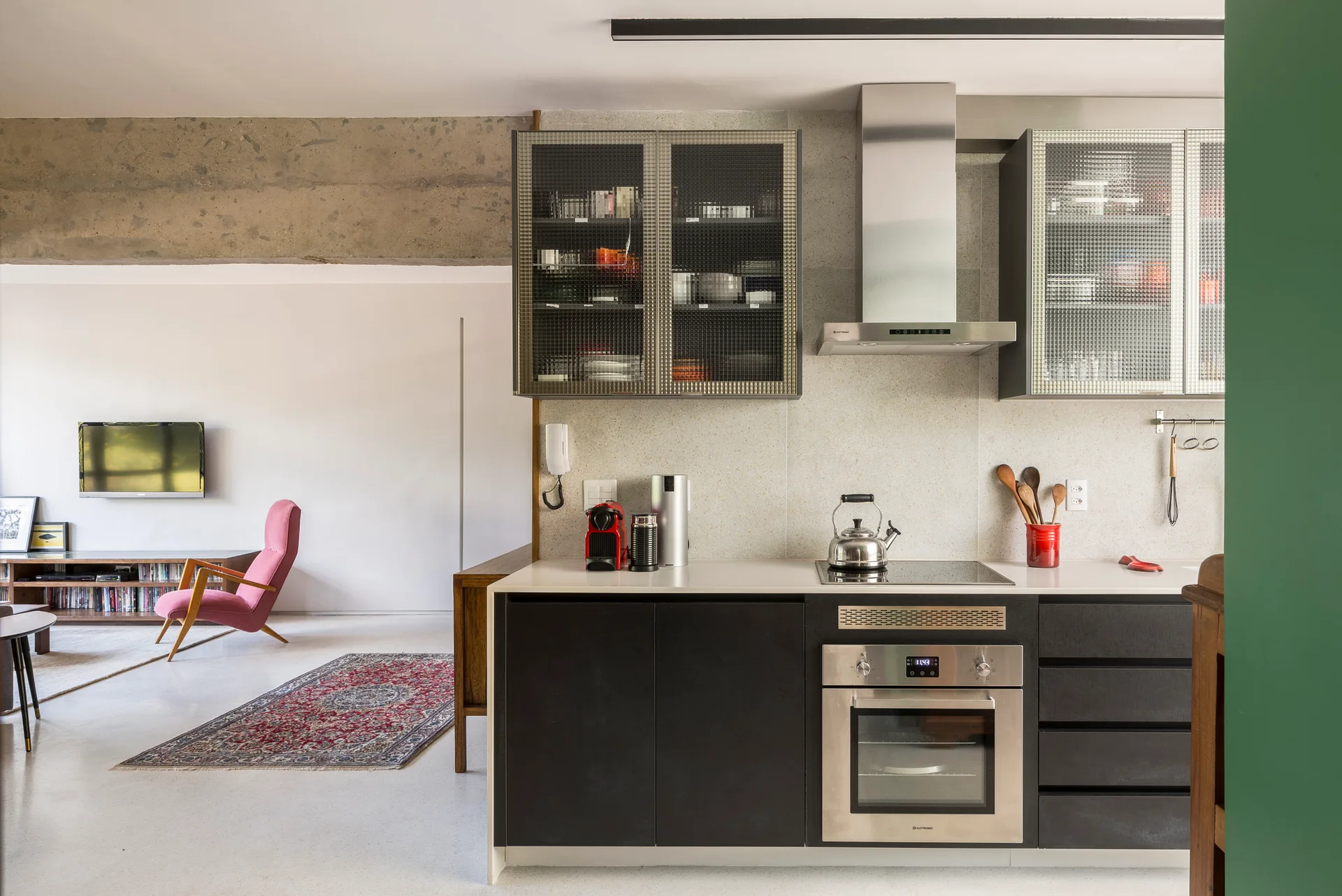 An open-plan kitchen features black cabinets, reeded glass doors, and a concrete beam looking into a living room with a pink chair.