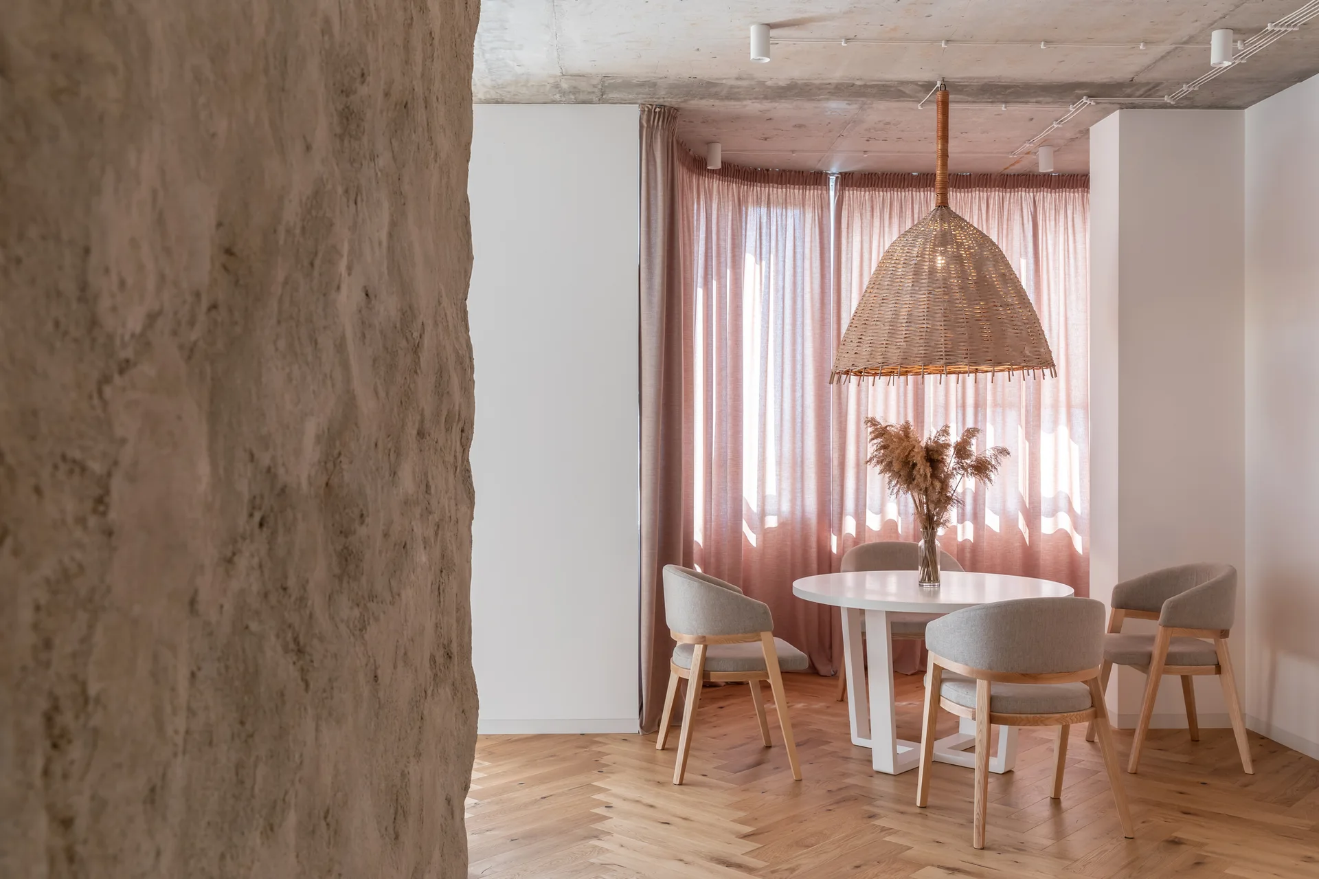 A round white dining table and grey upholstered chairs sit beneath a woven dome pendant light in front of dusty pink curtains.