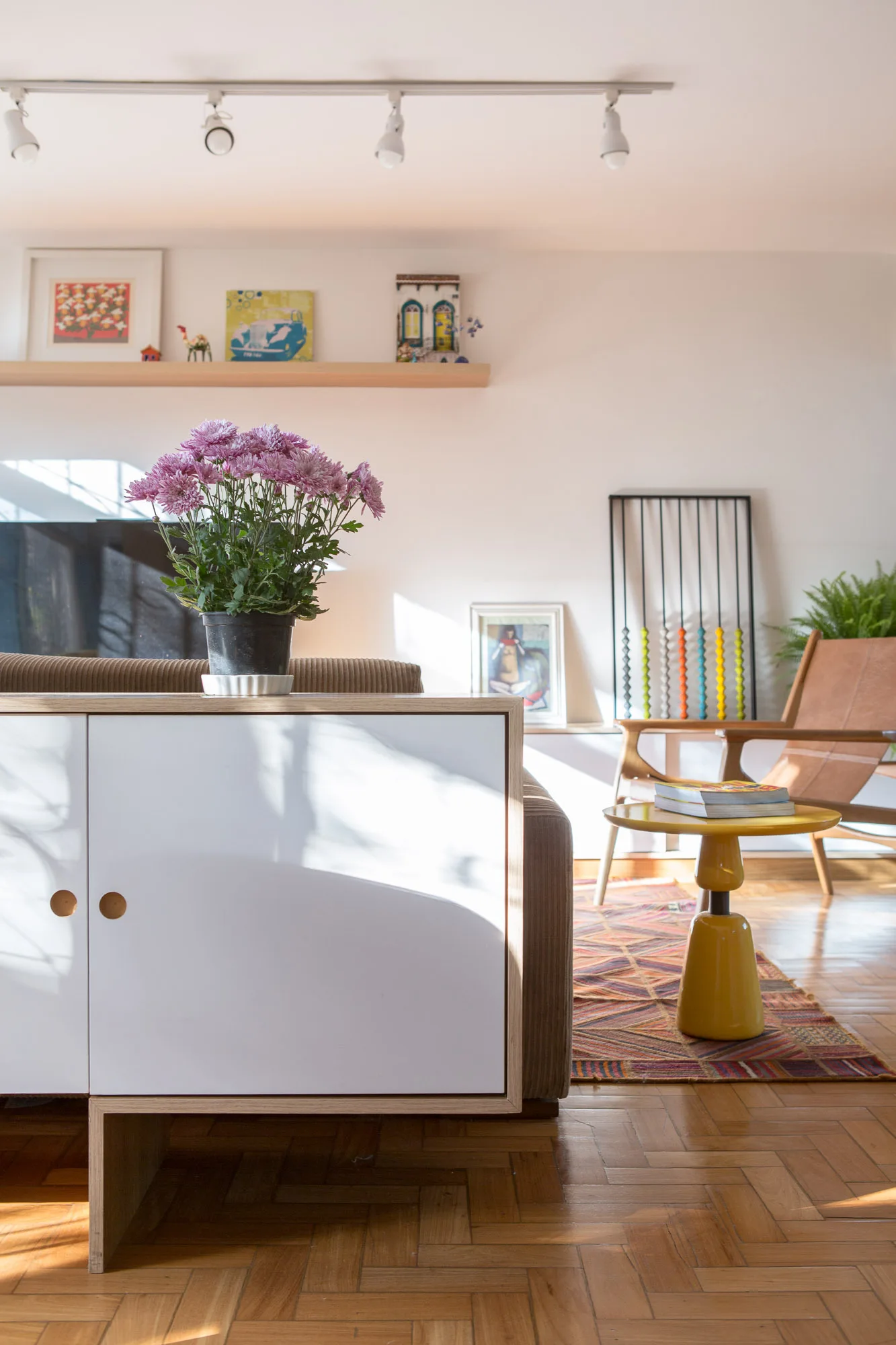 A living room features a white and wood sideboard, purple flowers, a yellow pedestal side table, and a leather sling chair on parquet flooring.