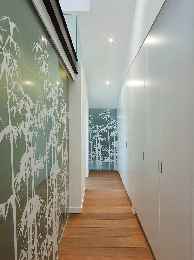A narrow hallway with hardwood floors features a seamless white storage wall and a green glass partition with a white bamboo motif.