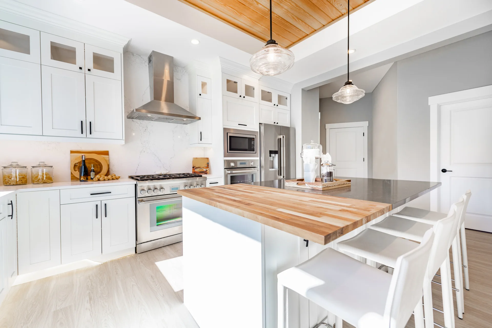 A bright kitchen features white shaker cabinets, stainless steel appliances, and a kitchen island with a wood countertop and white bar stools.