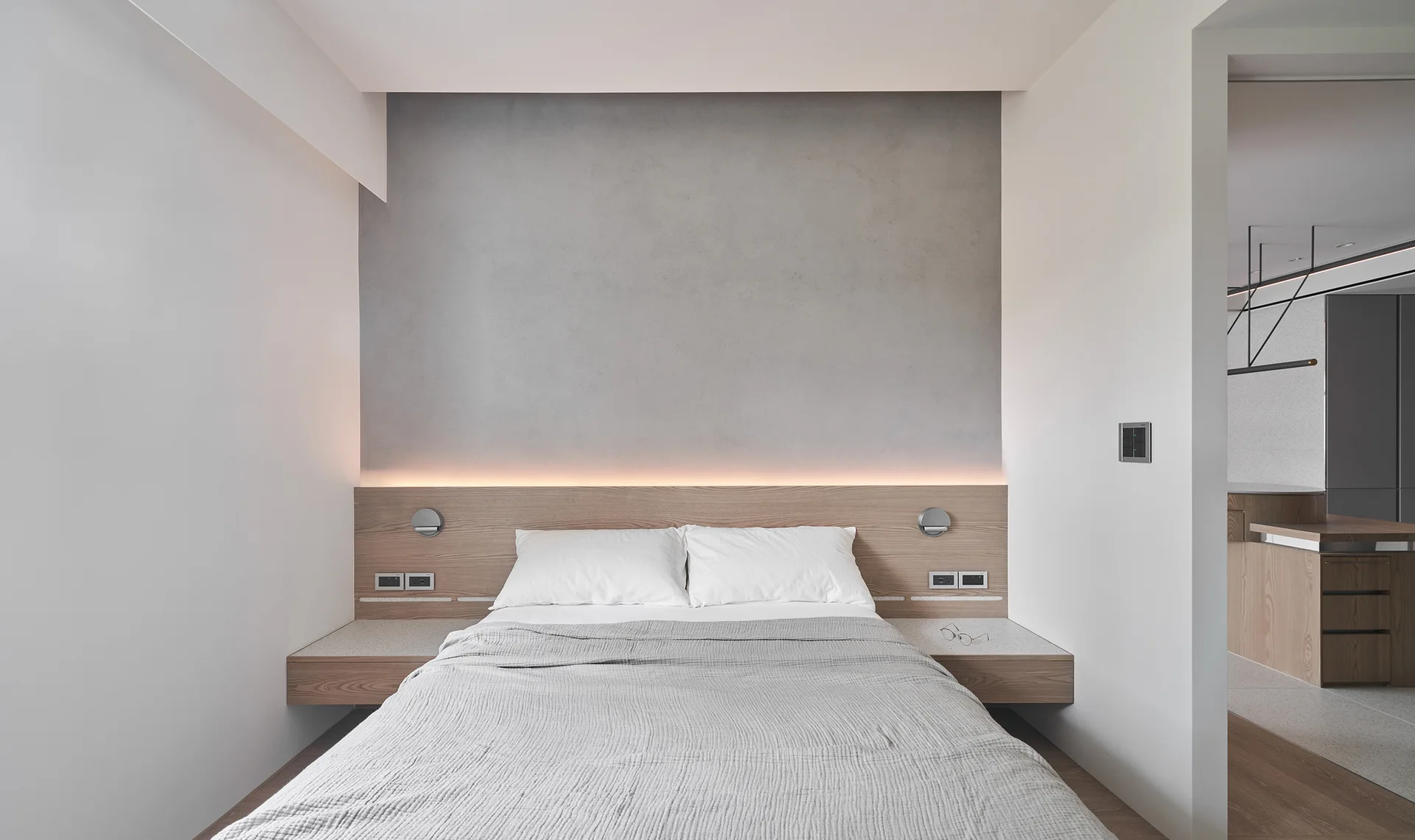 A minimalist bedroom features a bed with grey bedding, an integrated wood headboard with floating nightstands, and a textured grey wall with cove lighting.