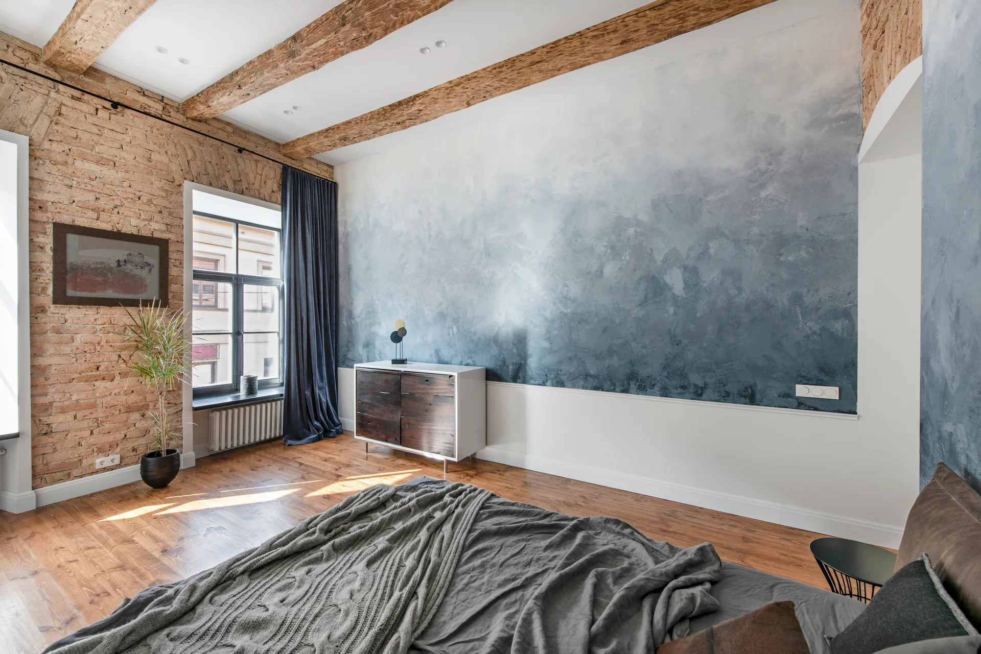 A bedroom featuring an industrial-style brick wall, exposed wooden ceiling beams, a blue gradient accent wall, and a bed with grey linens.