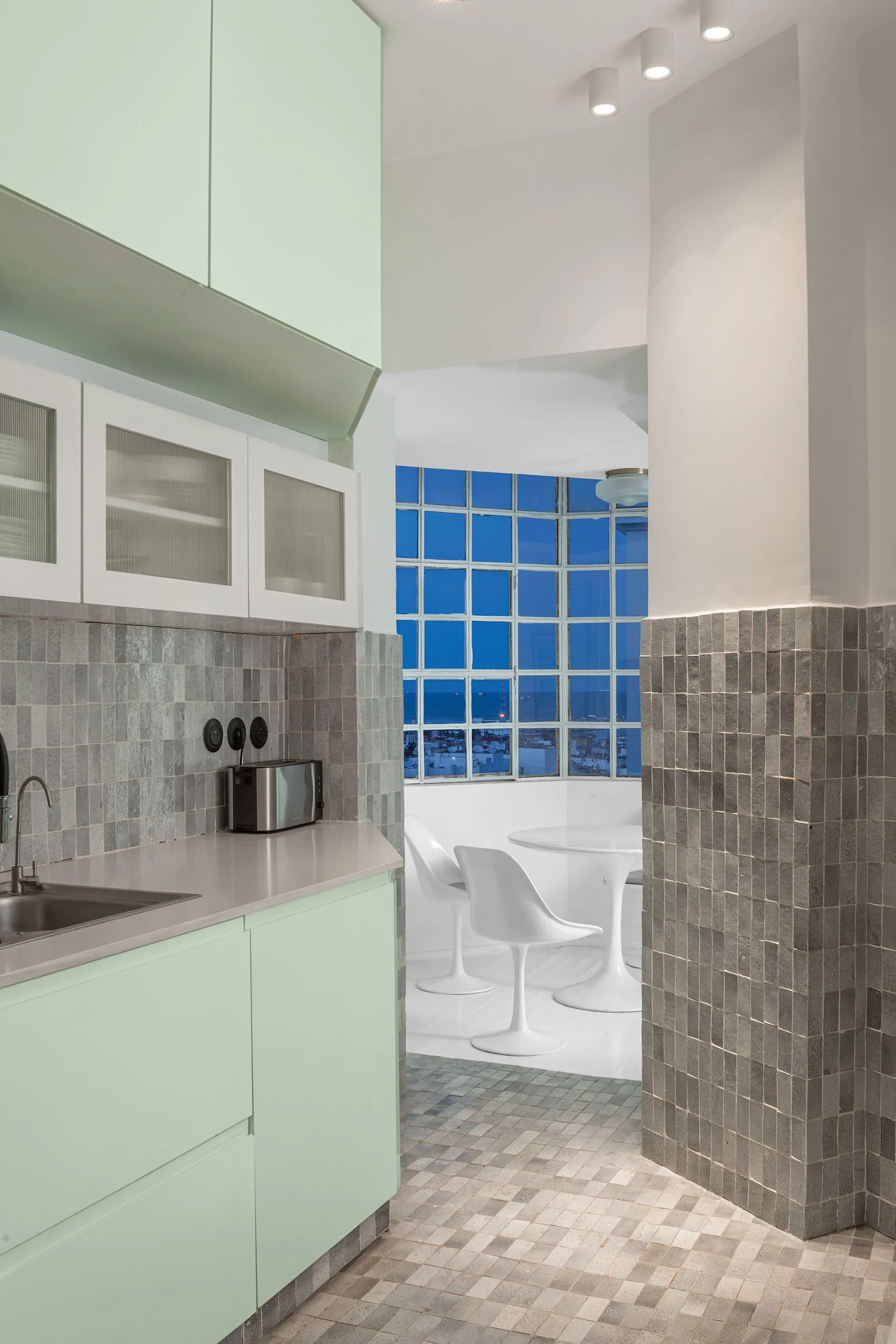 A modern kitchen with mint green cabinetry and grey square tile walls leading to a breakfast nook with a white pedestal table and chairs.