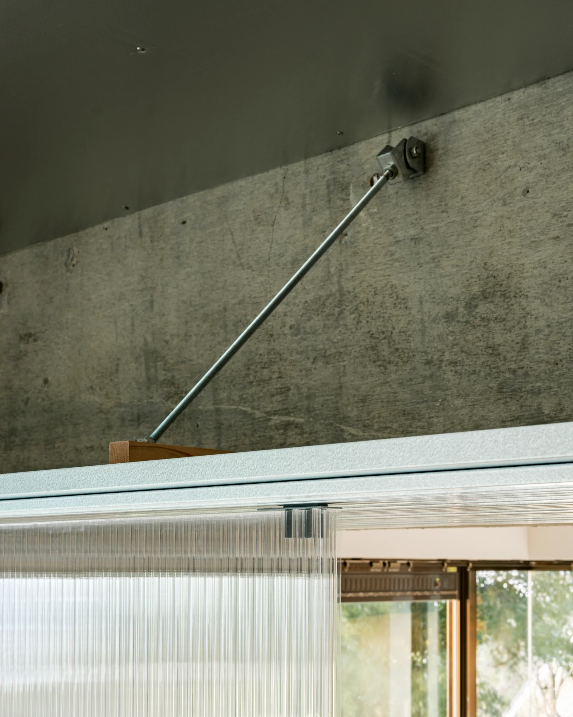 A metal tension rod anchors a translucent ribbed partition panel to a raw concrete wall.