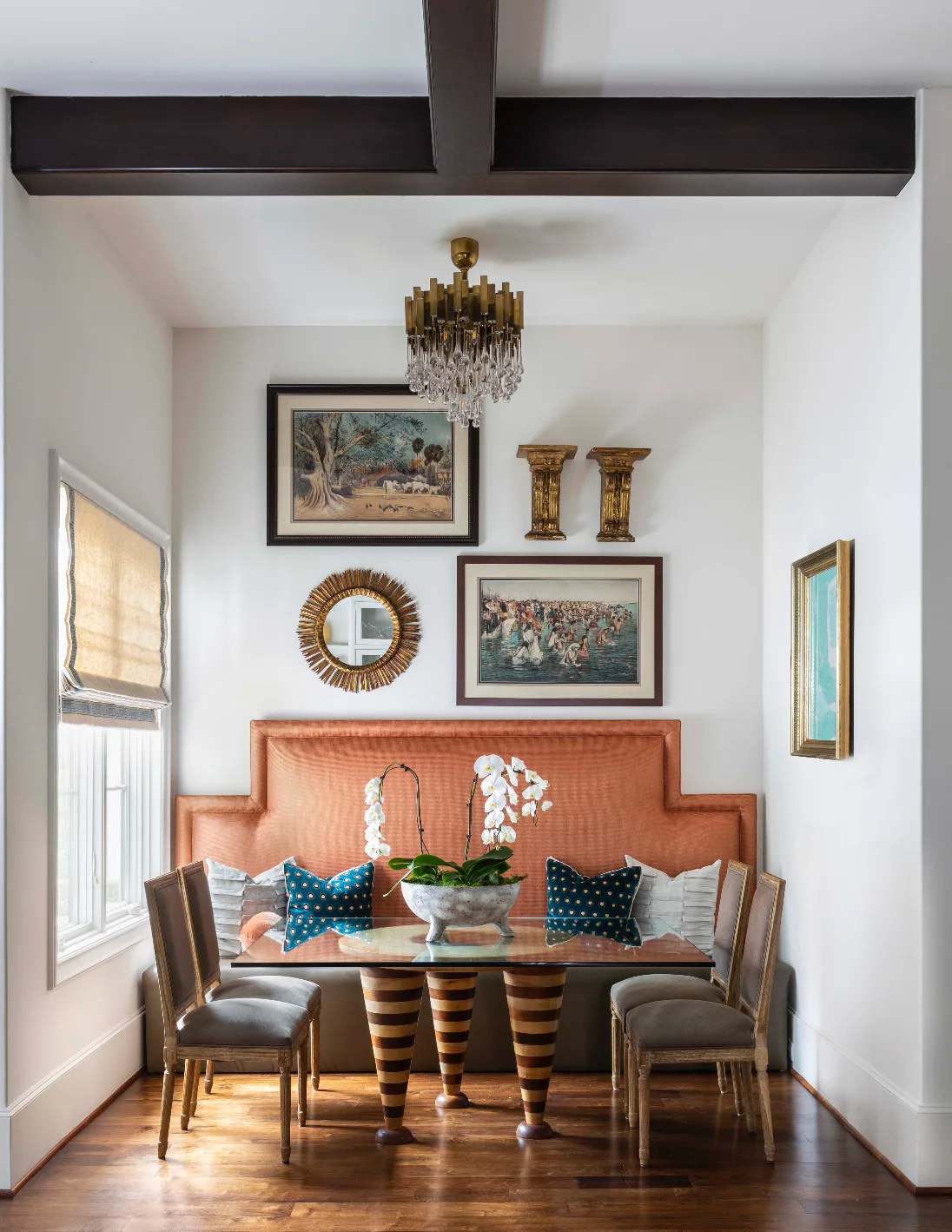 A dining nook featuring a tall orange banquette behind a glass-topped table with striped legs and chairs beneath a crystal chandelier.