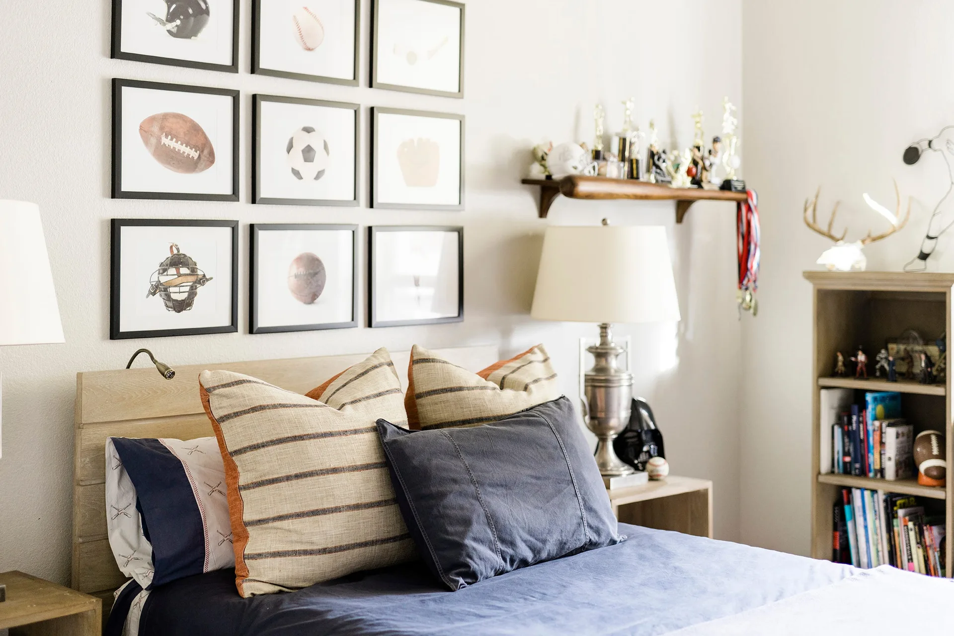 A child's bed with blue and striped pillows sits beneath a gallery wall of framed sports prints next to a wooden nightstand.