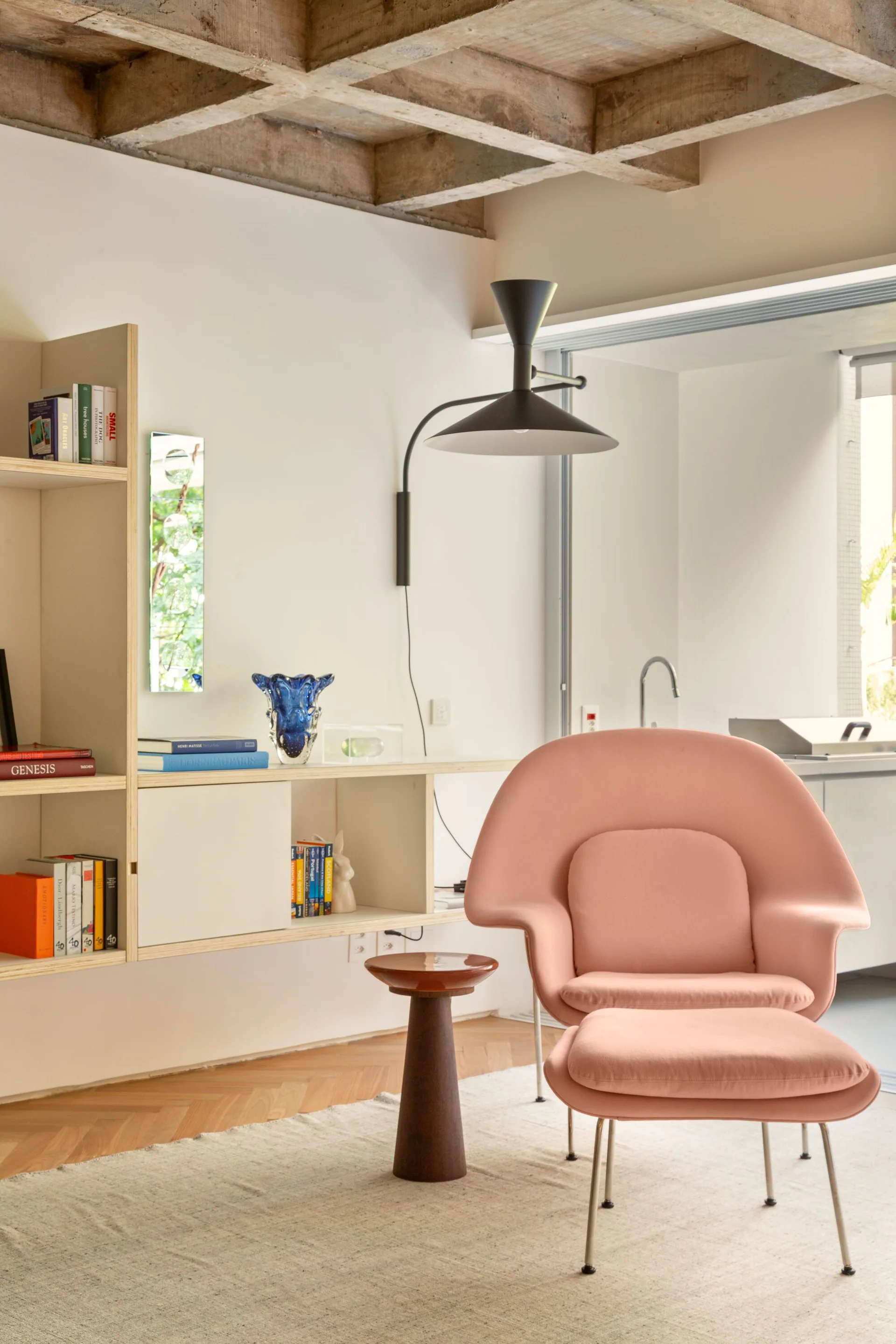 A pink lounge chair with an ottoman sits on a light area rug in a room with a light wood wall shelf and an exposed concrete grid ceiling.