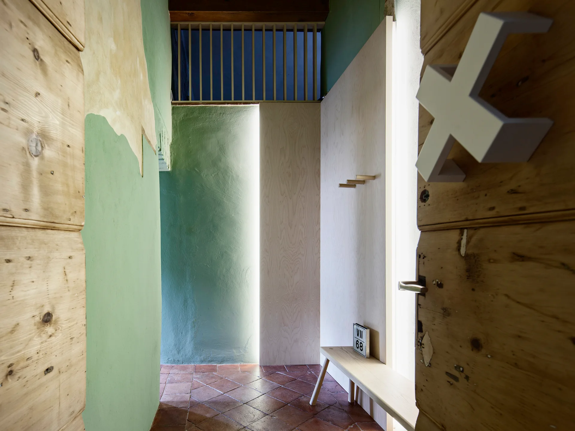 An entryway hallway featuring green plaster walls, diagonal terracotta floor tiles, and light plywood panels illuminated by vertical integrated lighting.