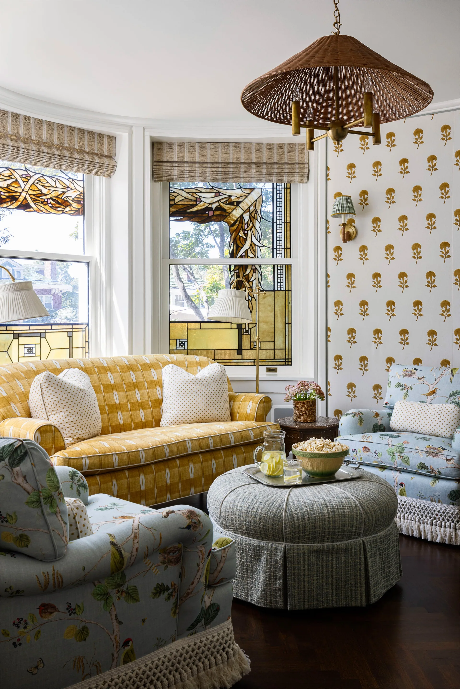 A cozy living room seating area features a yellow plaid sofa and floral armchairs arranged around a gray skirted ottoman by a stained glass bay window.