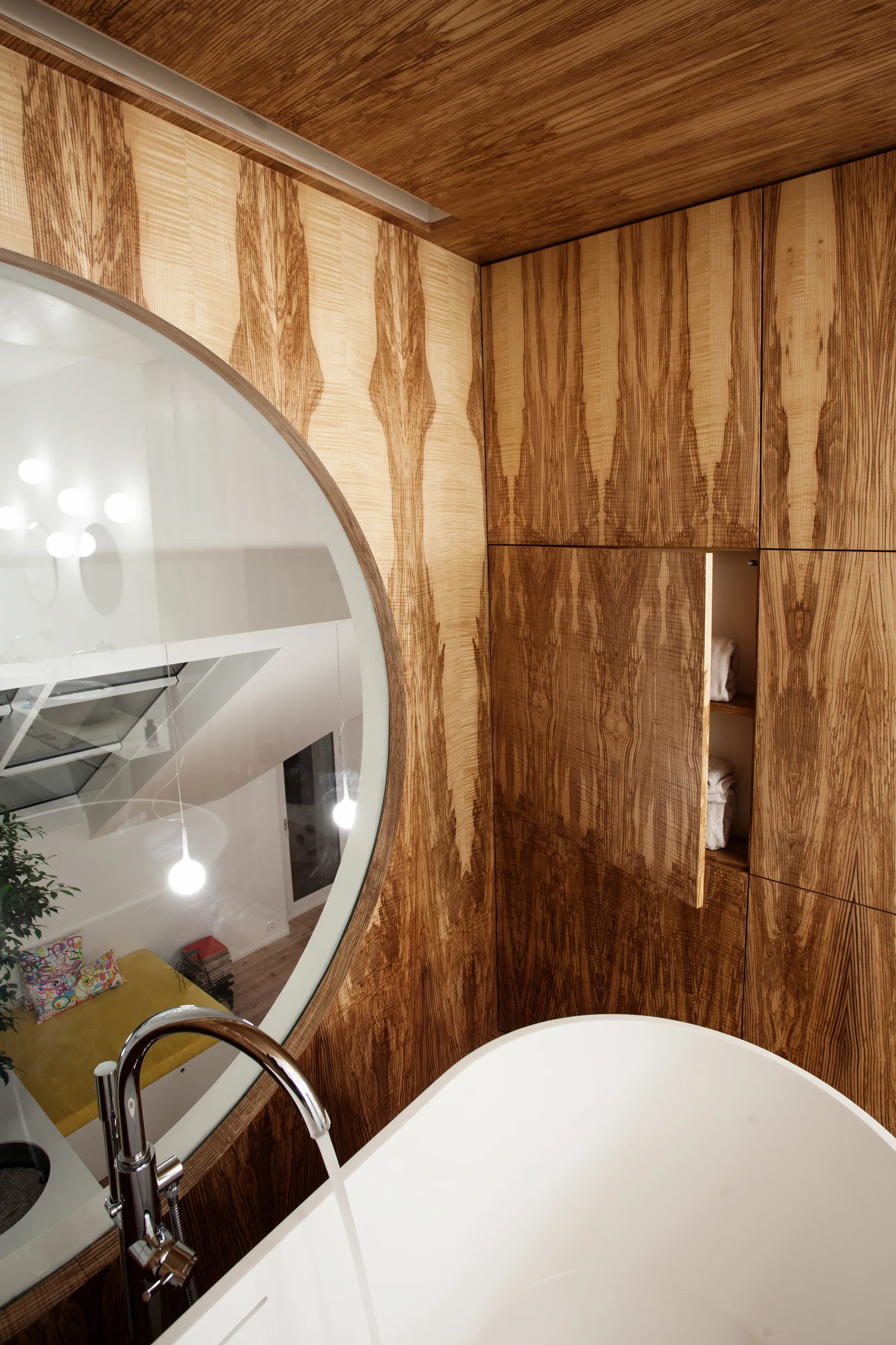 A white bathtub with a chrome faucet sits beside walls and cabinets covered in warm wood veneer next to a large circular wall mirror.