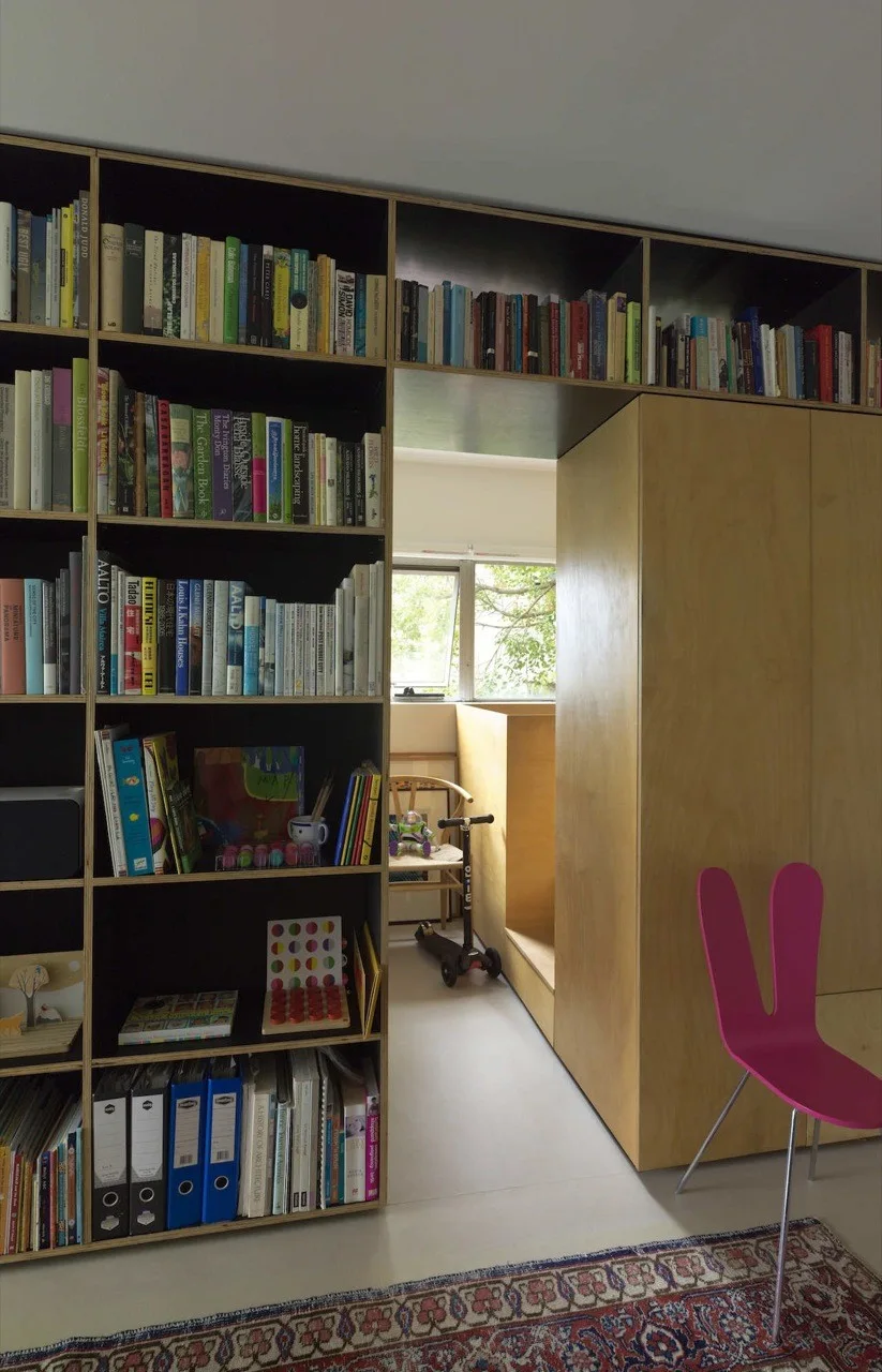 A custom plywood bookshelf wall built around a doorway features exposed edges and integrated storage, with a pink chair in the foreground.