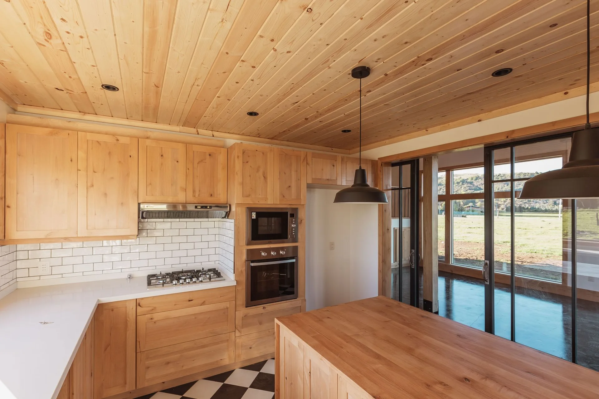 A kitchen with light wood cabinets, matching wood ceiling, white tiled backsplash, black pendant lights, and a black and white checkered floor.