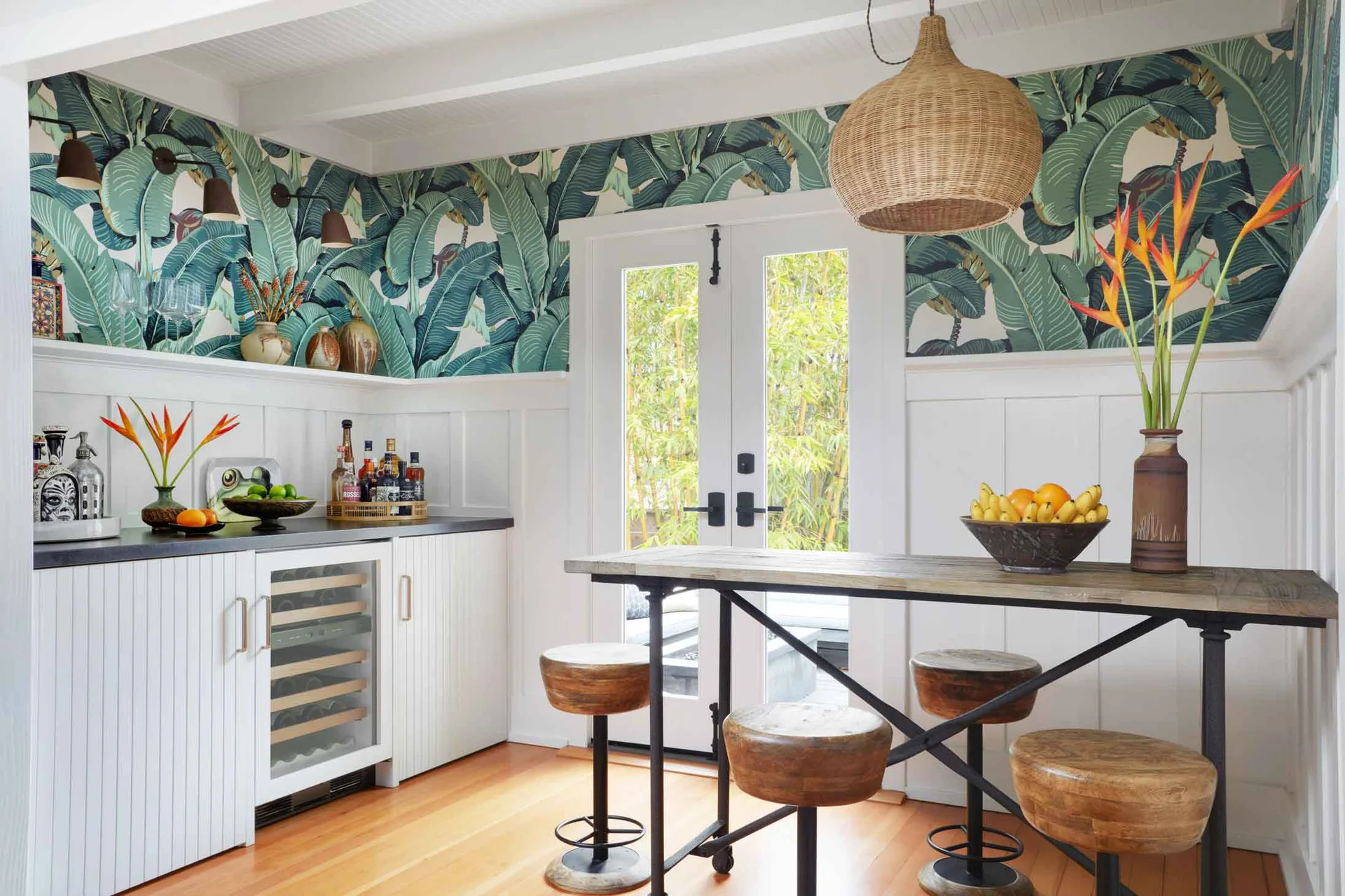 A wet bar area features green tropical leaf wallpaper, white wainscoting, an undercounter wine fridge, and a tall wood bar table with round stools.