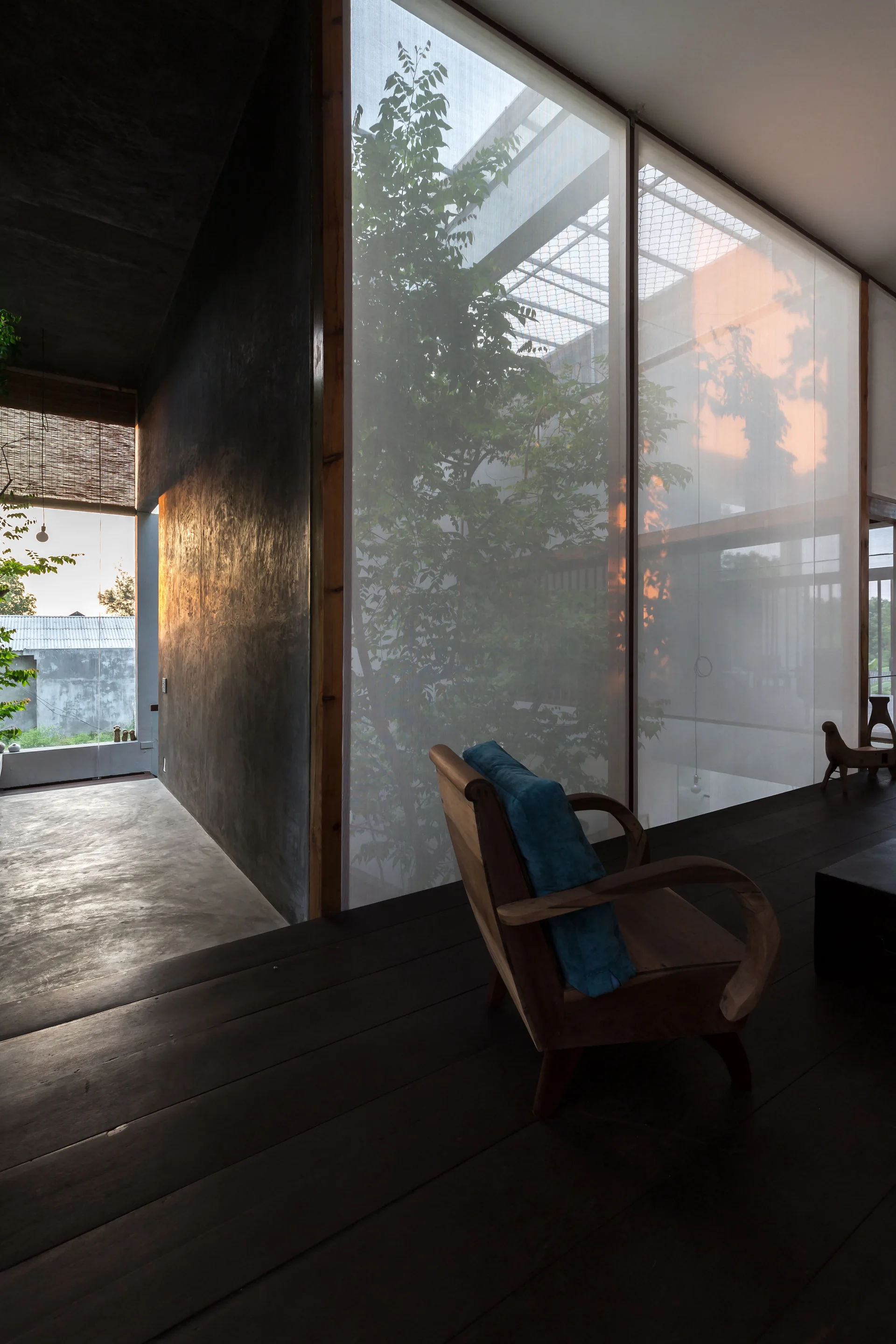 A dark wooden armchair sits on dark timber floors before a large floor-to-ceiling glass wall covered in a translucent white screen with trees behind it.