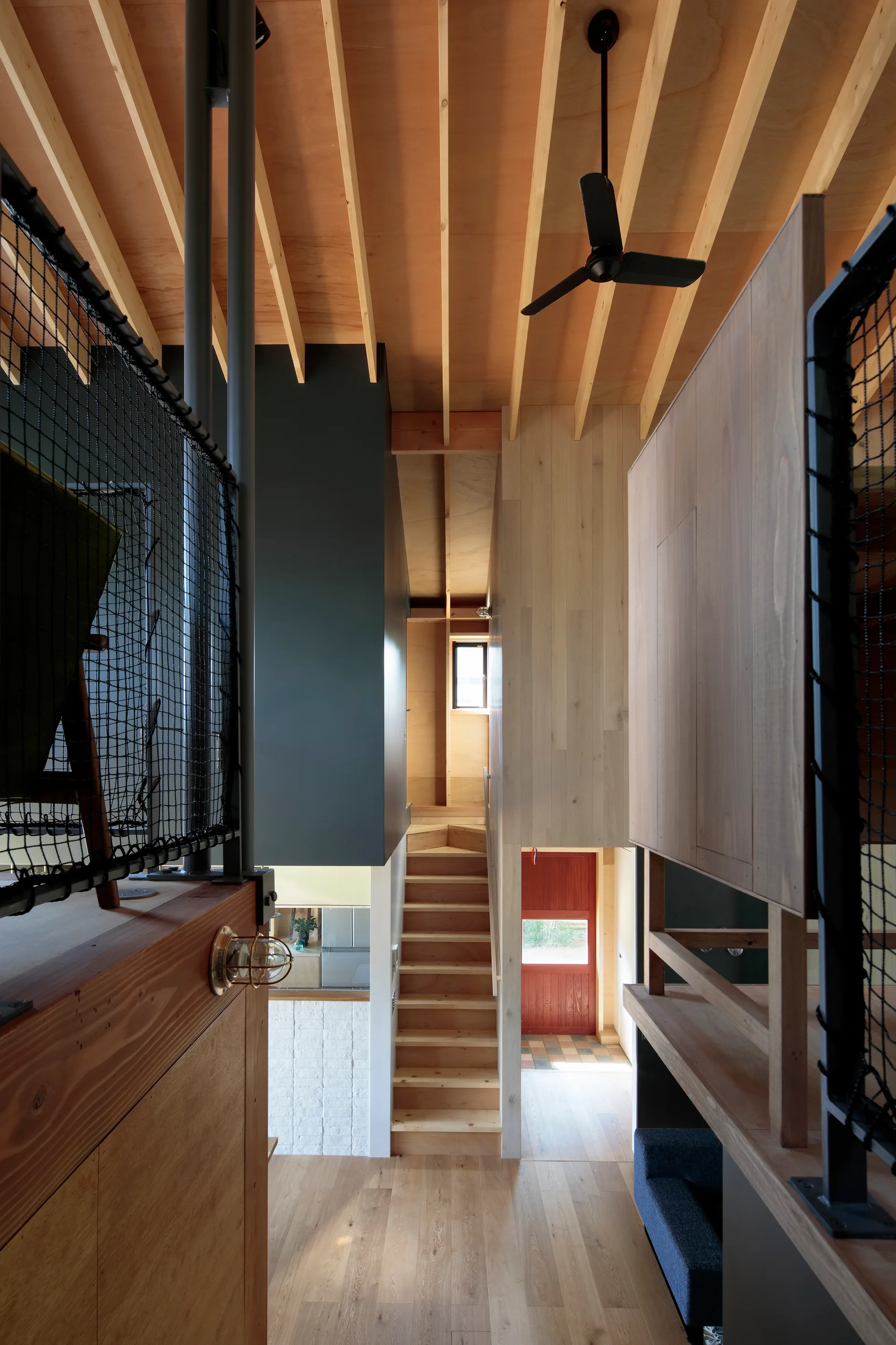 A modern interior residential wooden staircase with a black mesh safety railing and exposed ceiling beams leading to a red door in the background.