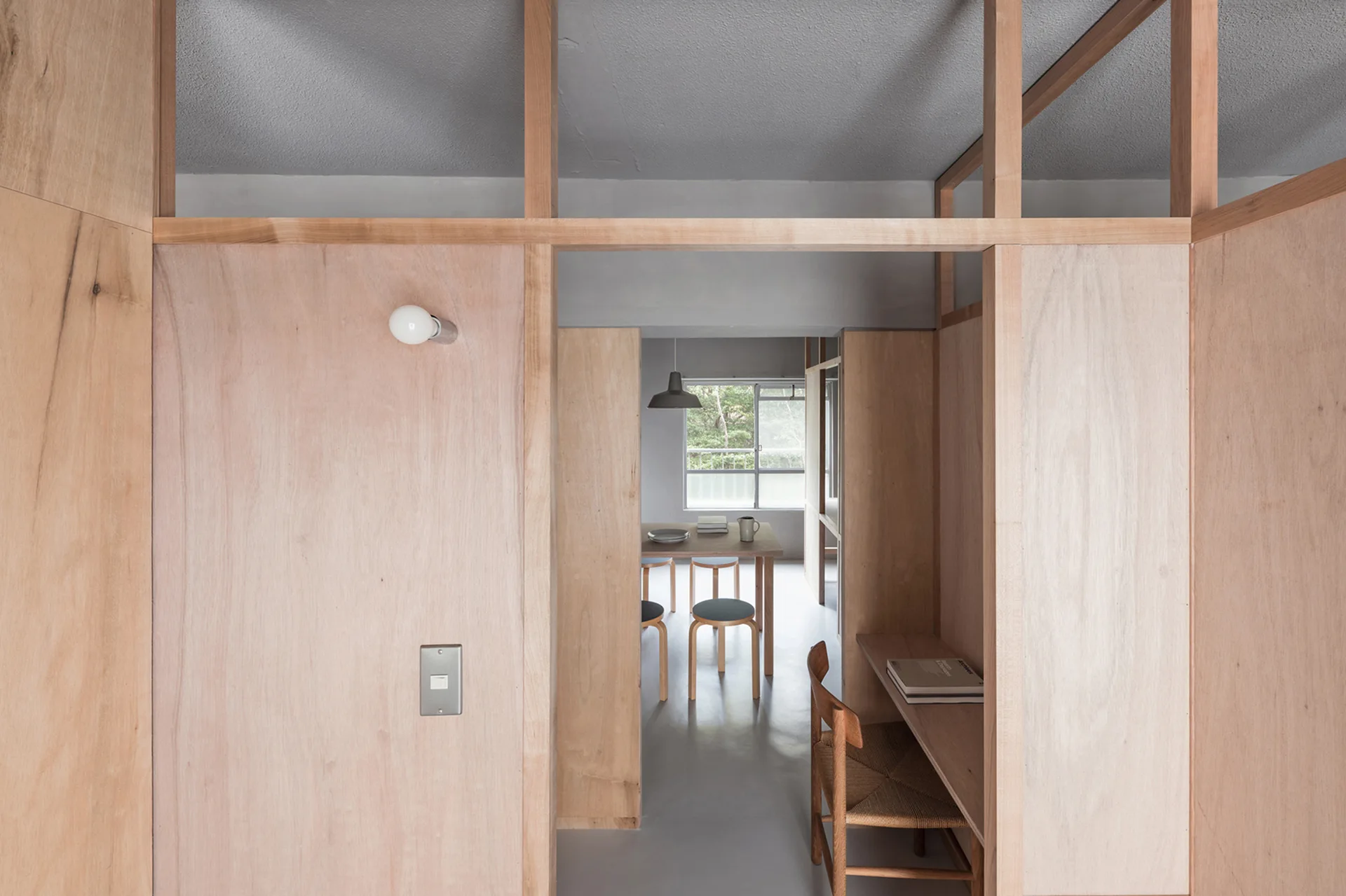 A light wood plywood partition wall with a built-in desk opens into a dining area featuring a wooden table and chairs.