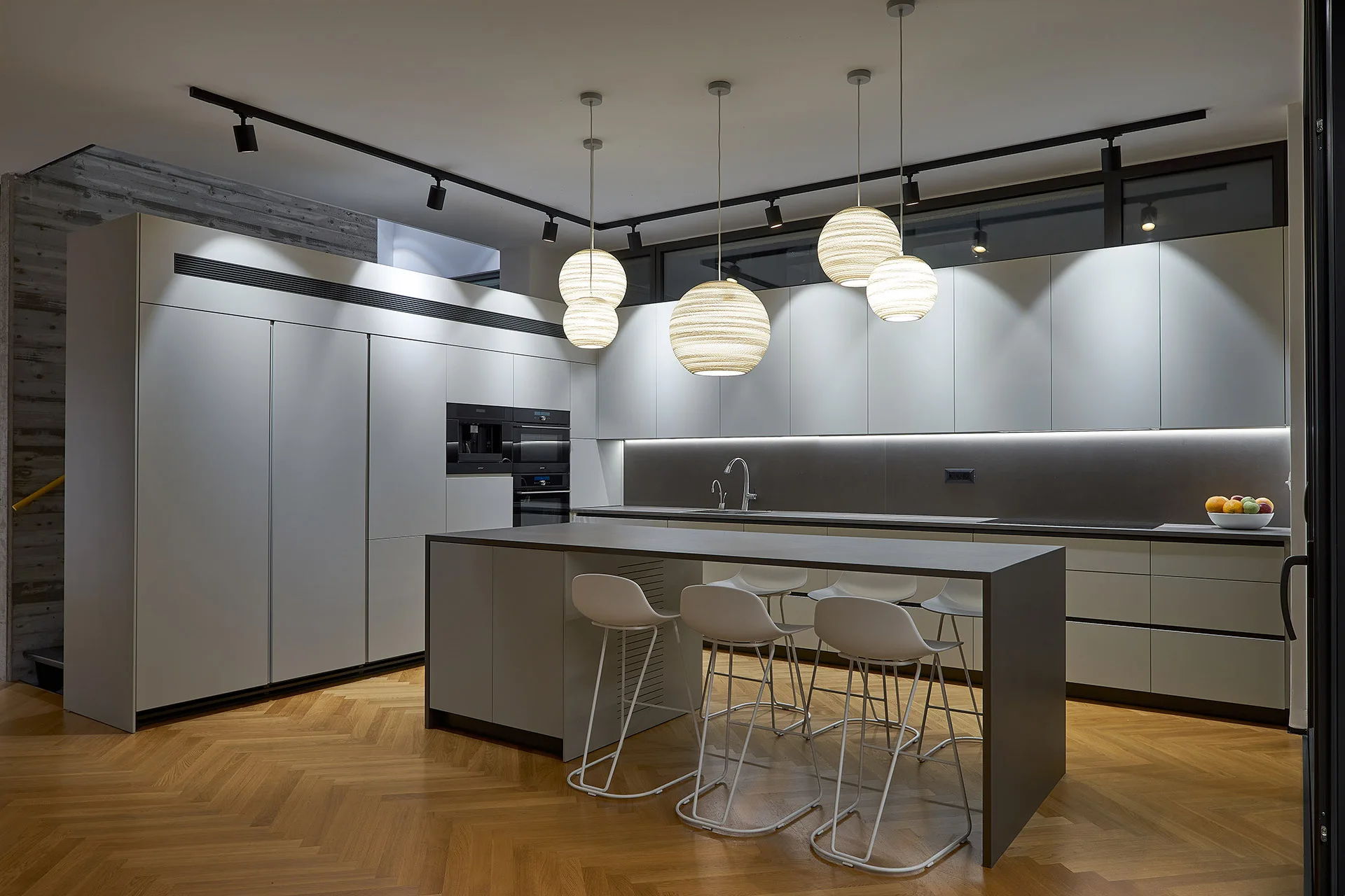 A modern gray kitchen features handleless cabinets, a central island with white bar stools, herringbone wood floors, and hanging spherical pendant lights.
