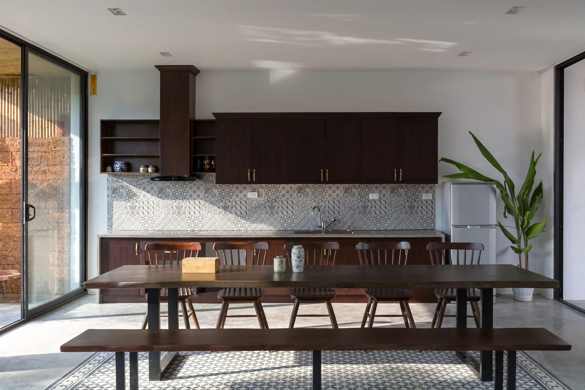 A dining kitchen with dark wood cabinetry, a large matching wooden dining table and bench, patterned tile backsplash, and concrete floors.