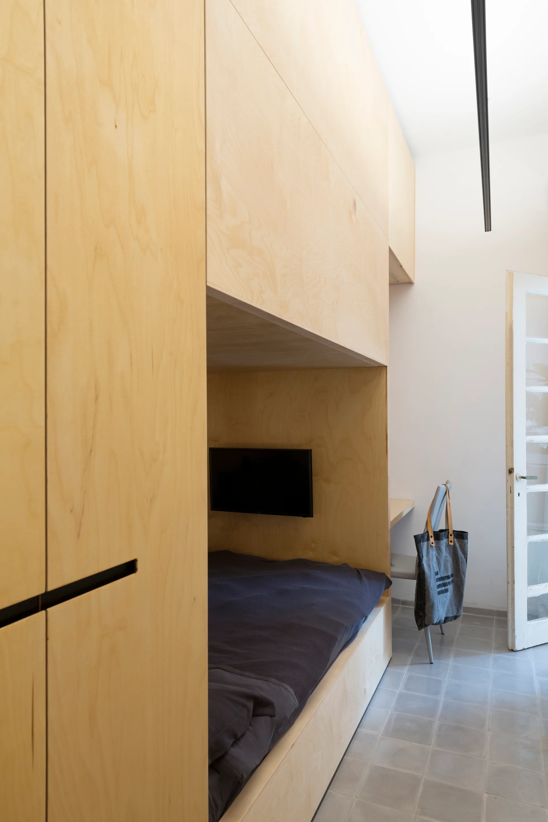 A custom light plywood storage cabinet unit features an integrated sleeping nook with dark blue bedding and a wall-mounted television.