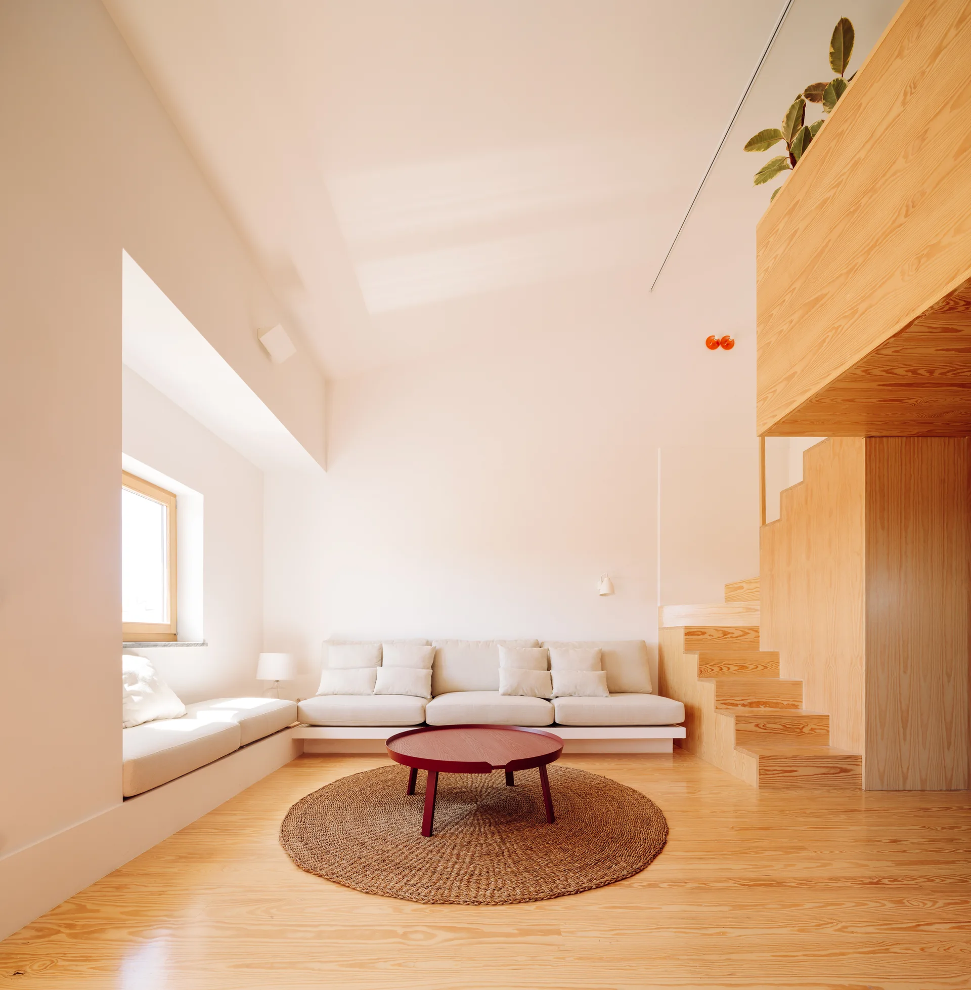 A bright minimalist living room features an off-white sofa, a custom light-wood staircase, a window bench, and a round red coffee table on a woven rug.