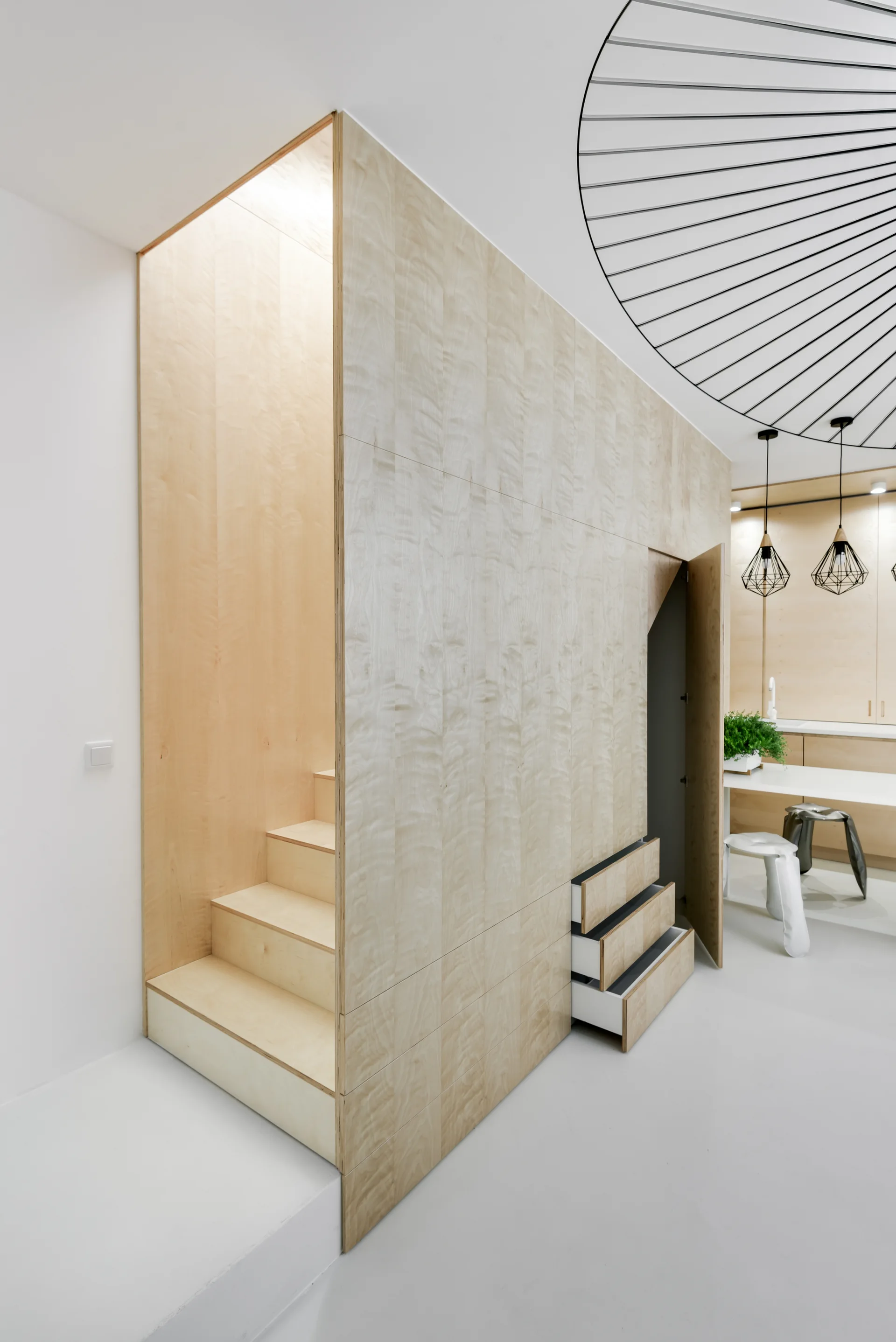 A custom light plywood staircase features integrated pull-out drawers and a closet door in a bright, minimalist apartment.