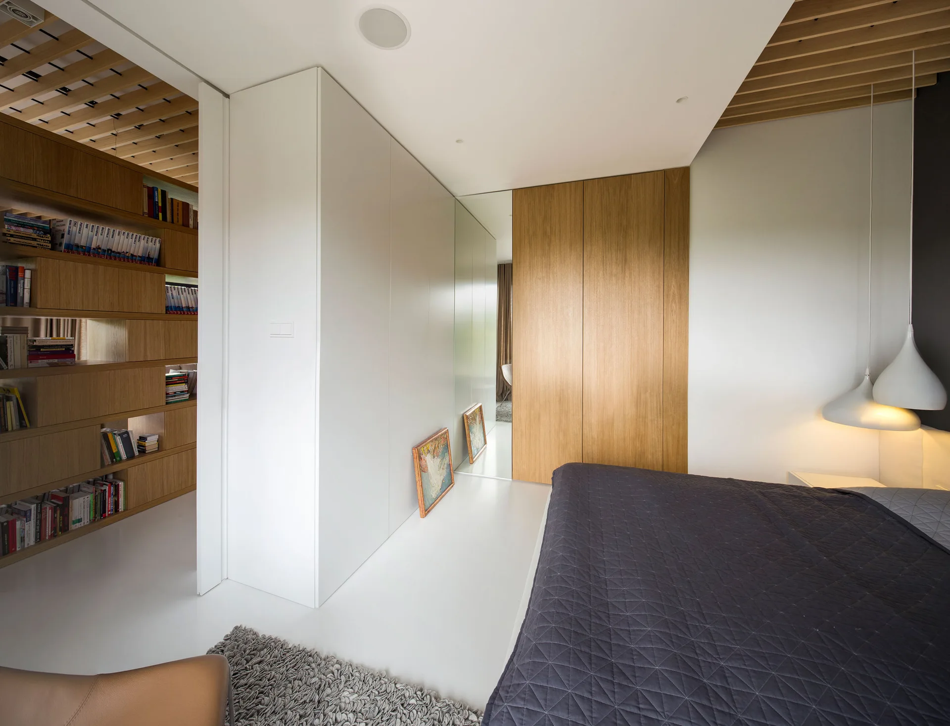 A modern bedroom features a grey quilted bed, white and wood built-in wardrobes with a mirror panel, and a wood slat ceiling.