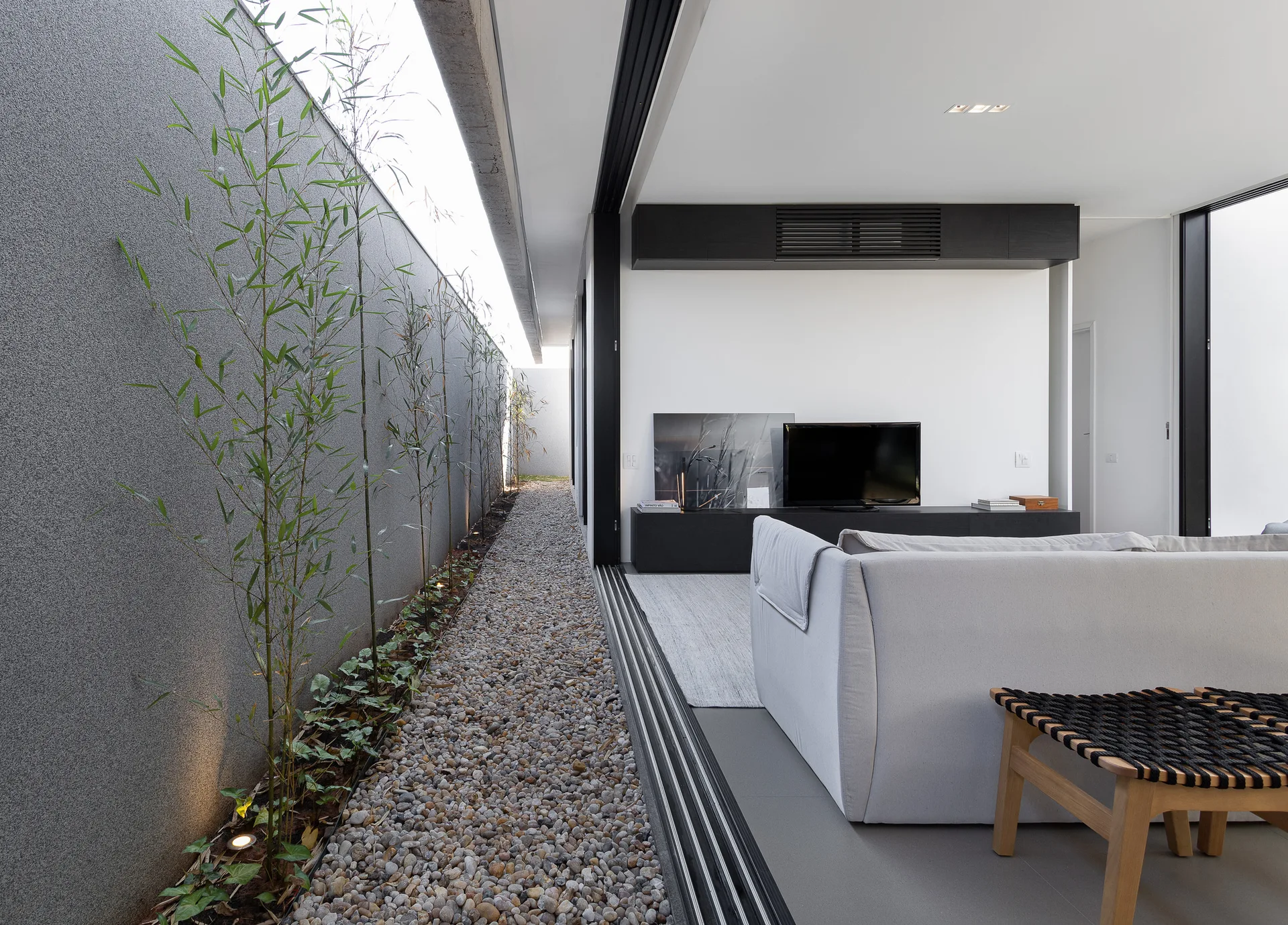 A modern living room with a grey sofa and TV console opens through sliding glass doors to a narrow gravel courtyard planted with bamboo.
