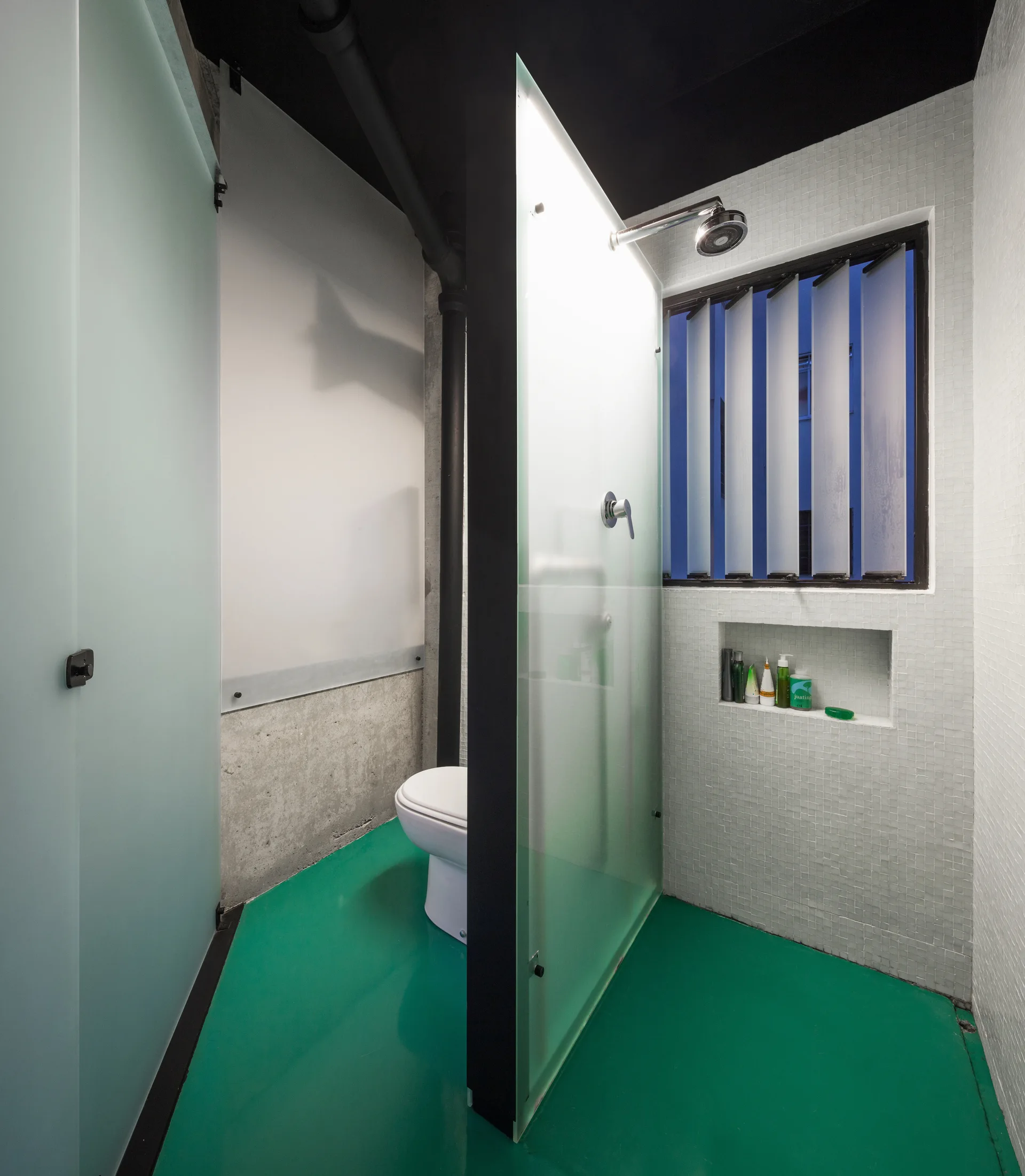 A modern bathroom with a glossy green floor, white tiled walls, a frosted glass partition, and an exposed black pipe near the ceiling.