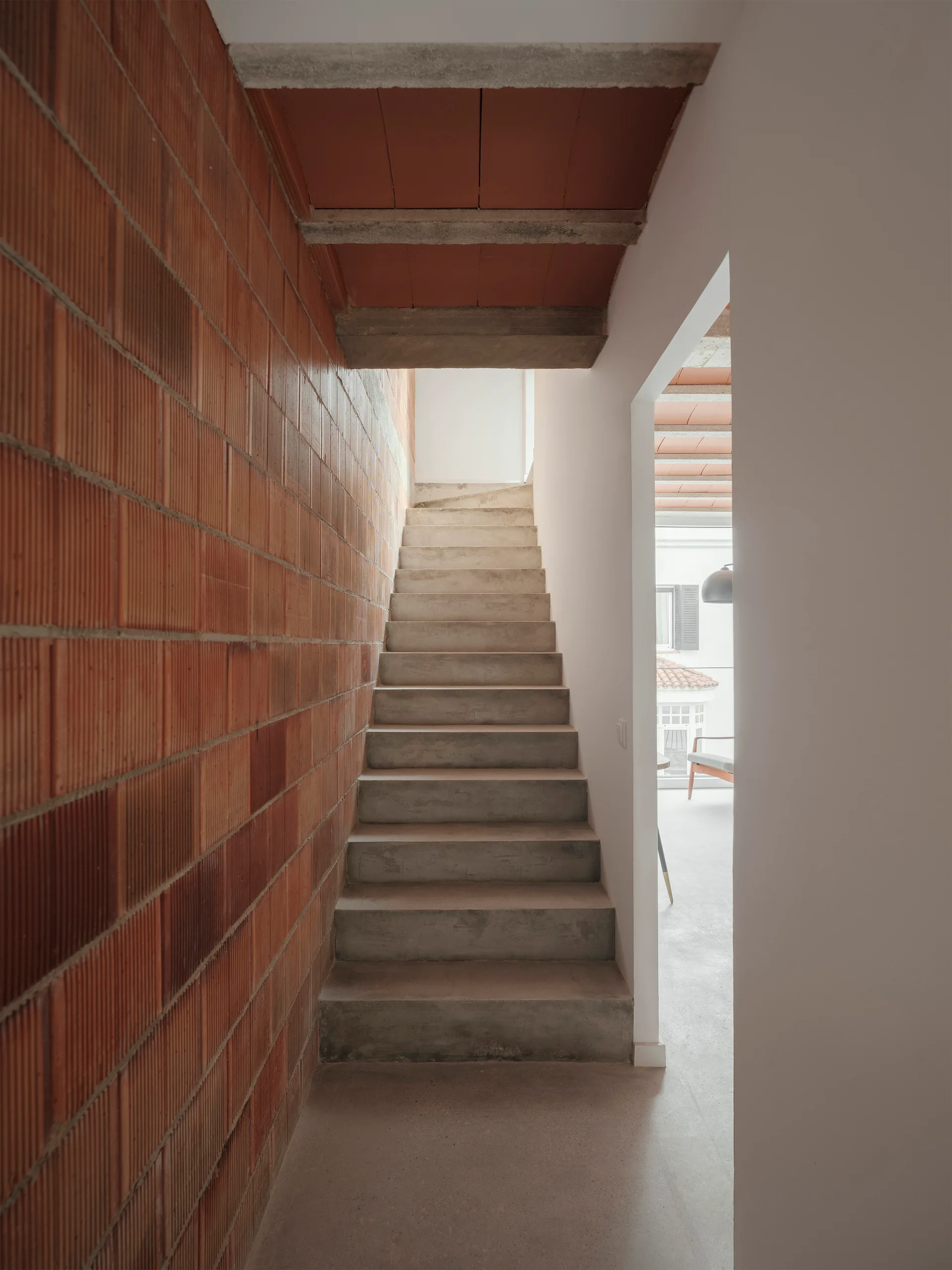 A straight concrete staircase rises between a textured terracotta brick wall and a smooth white plaster wall with a doorway.