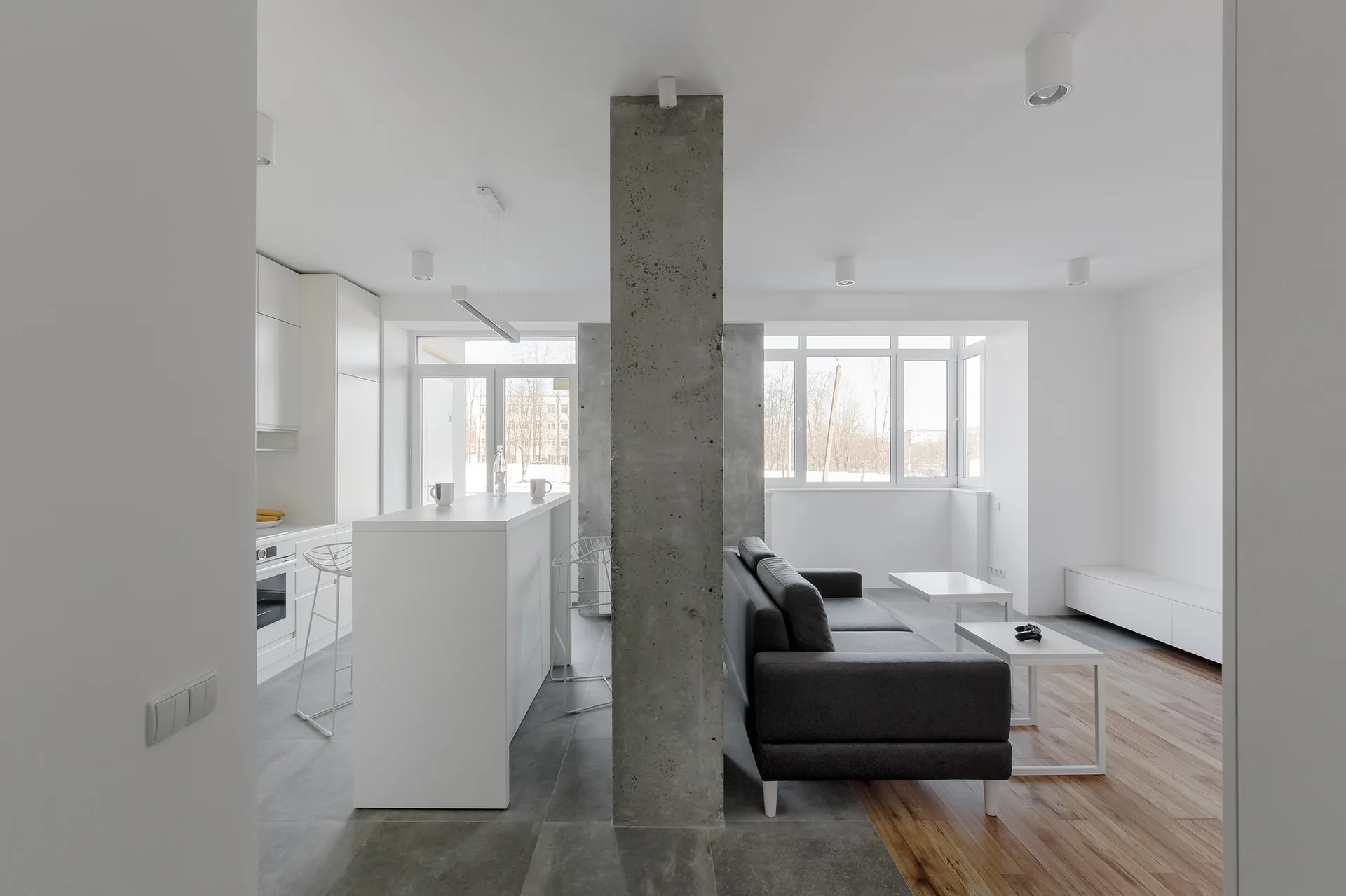 A minimalist open-plan living room and kitchen featuring a central raw concrete pillar, a dark grey sofa, and split tile and wood flooring.