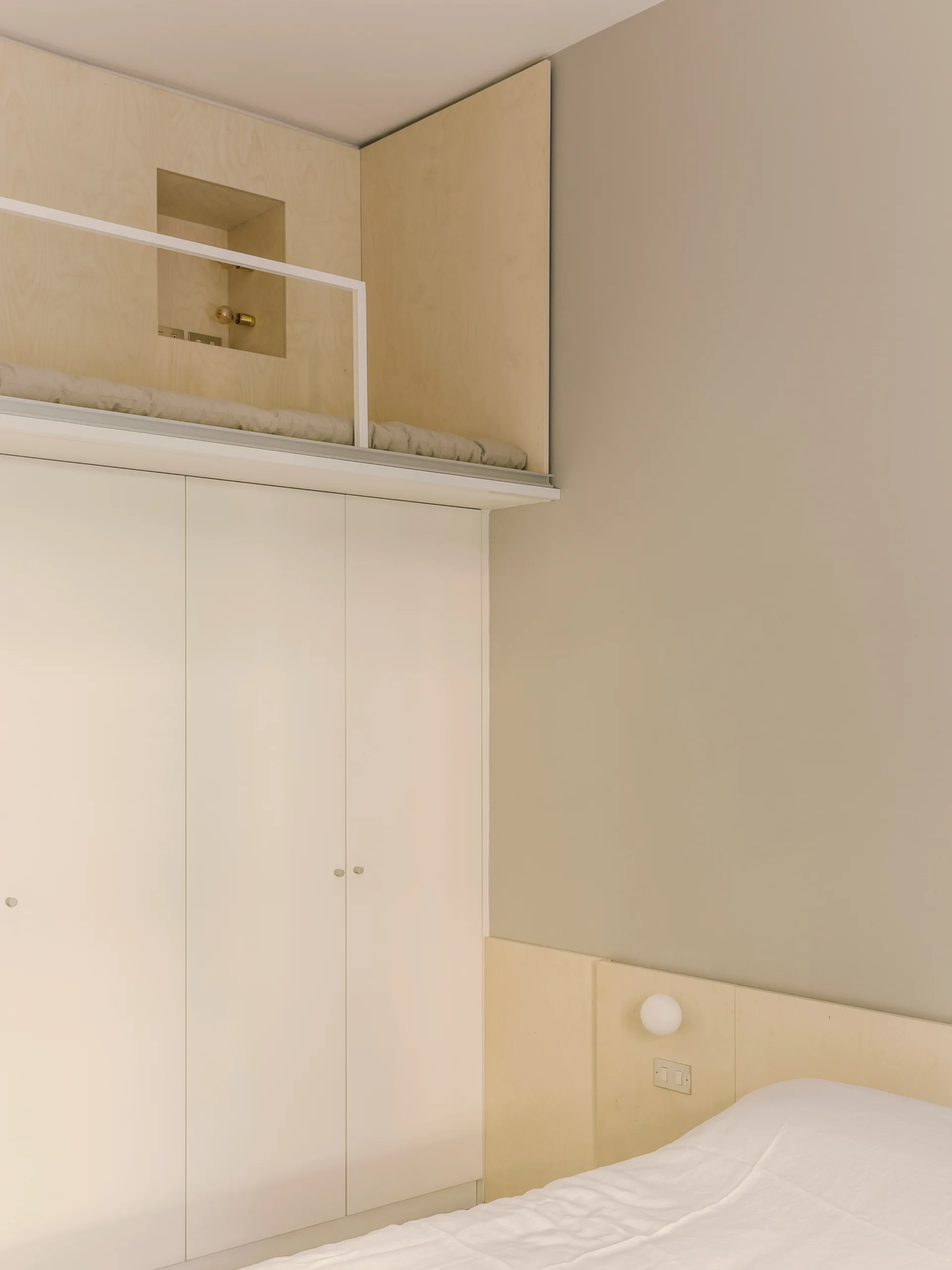 A minimalist bedroom featuring a custom light wood loft above white built-in wardrobes, set against a warm beige wall with a lower bed.