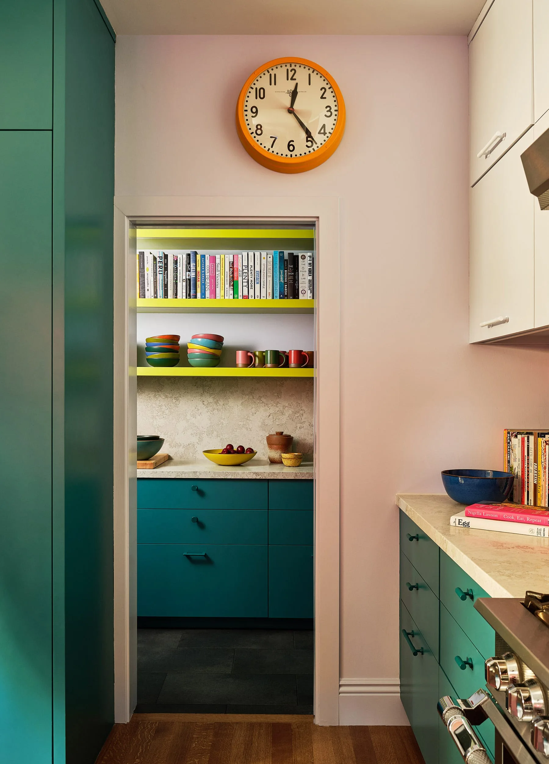 A kitchen doorway opens to a pantry with teal cabinets, illuminated bright green shelves holding cookbooks, and a large orange clock on the pink wall above.