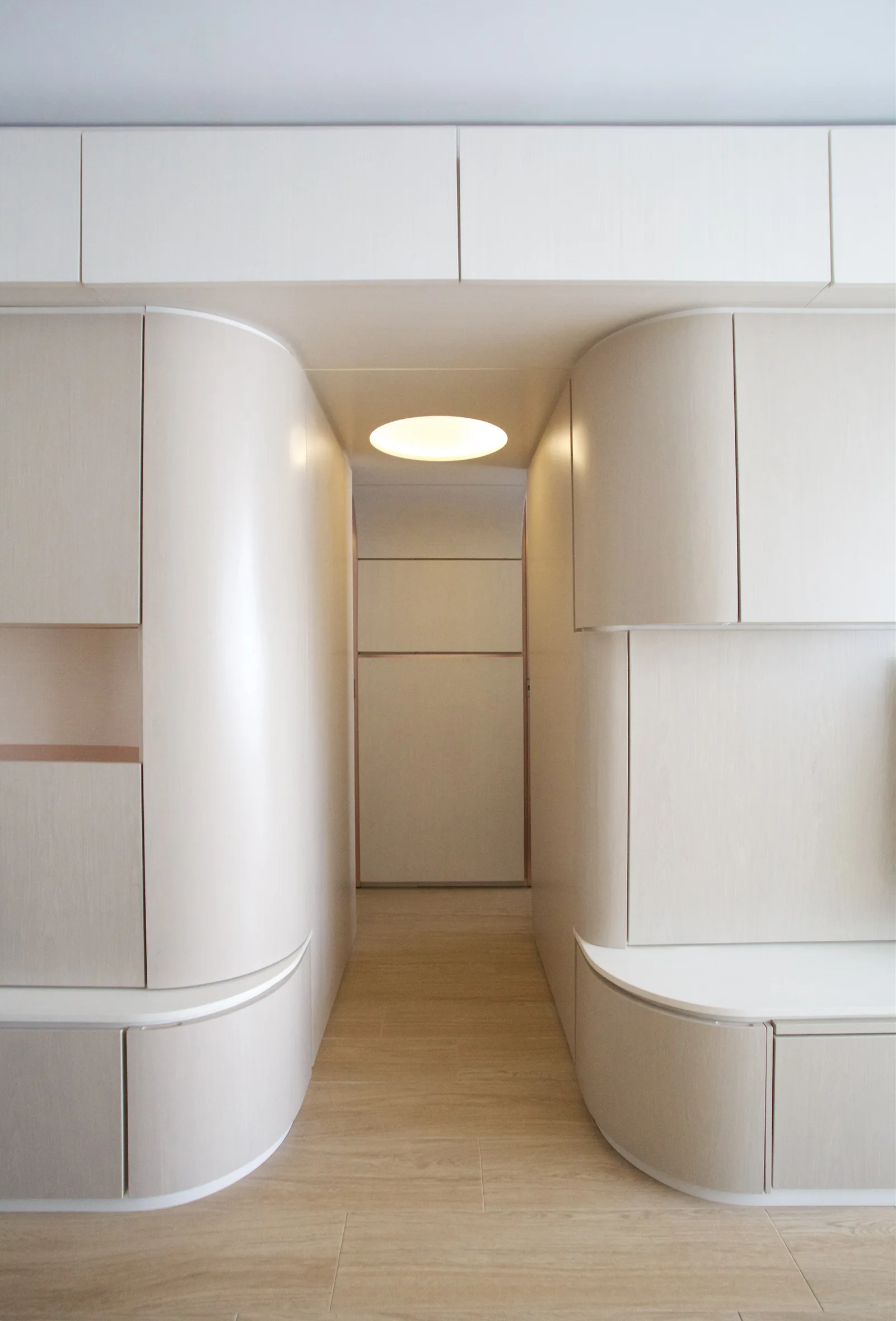 A narrow hallway features seamless light wood-toned built-in cabinets with curved corners and a recessed circular light in the ceiling.