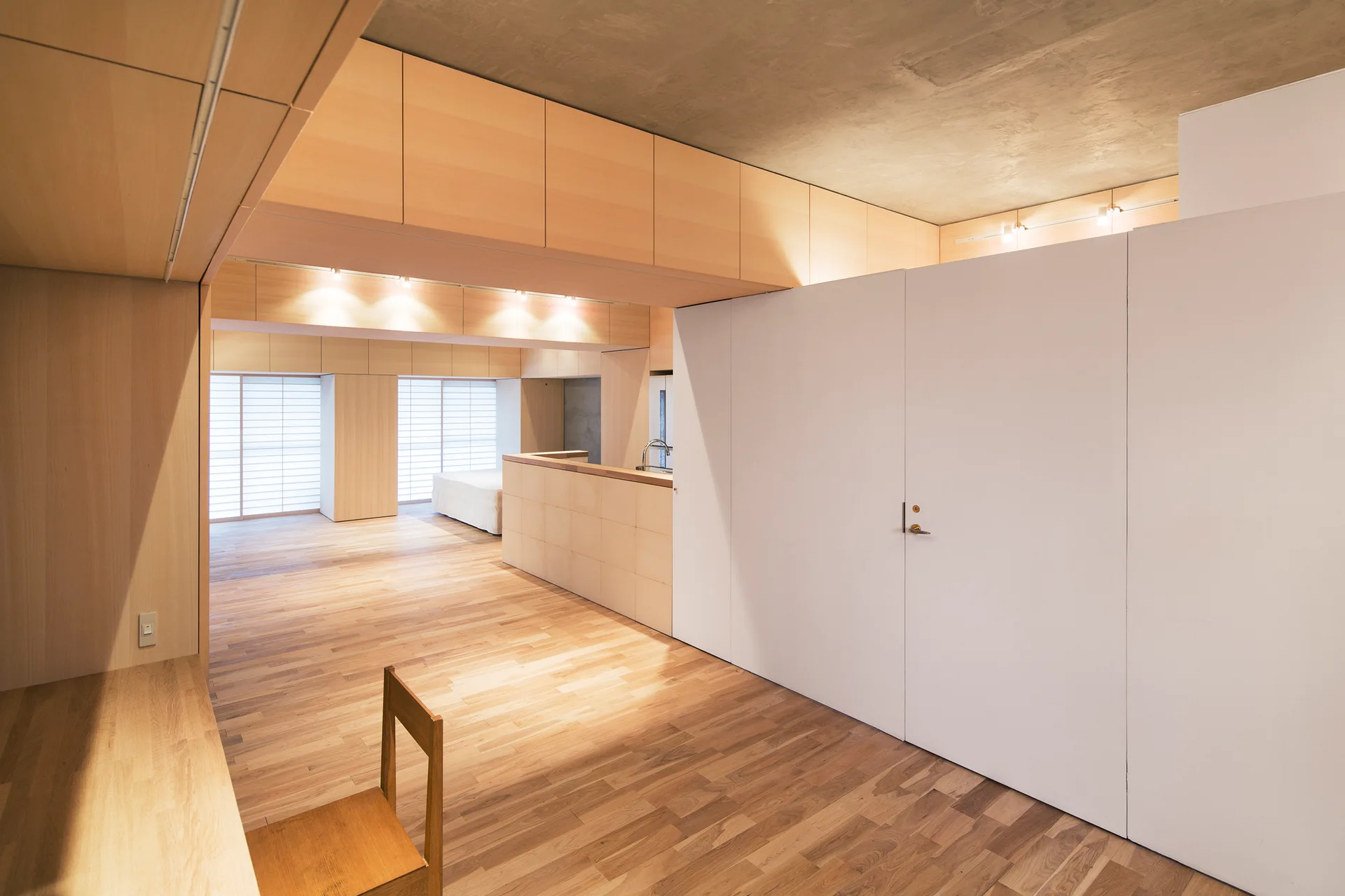 A minimalist open-plan room features light wood flooring, integrated wood cabinetry, a large white partition wall, and translucent shoji screens.