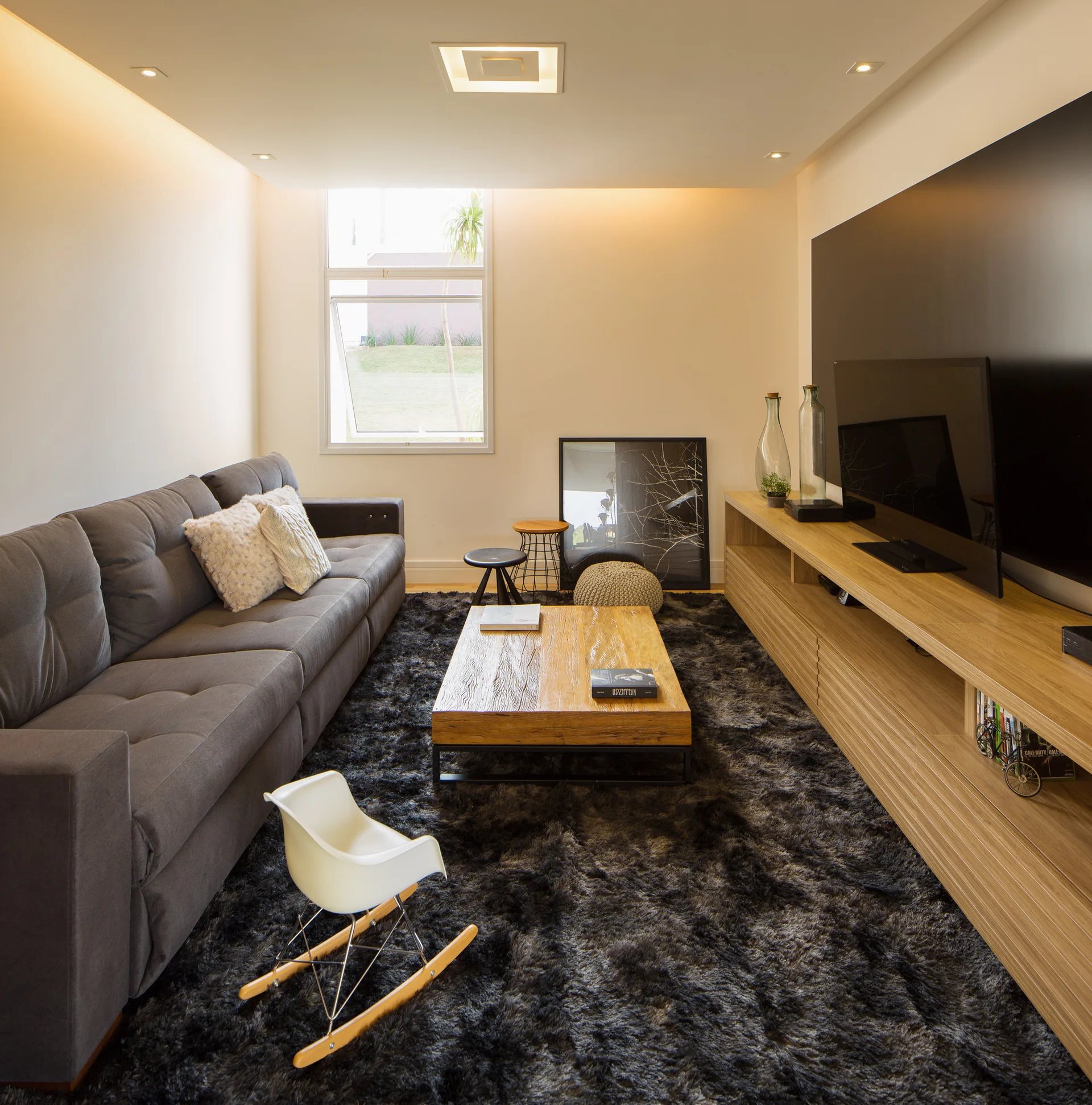 A contemporary living room featuring a grey tufted sofa, a long wood media console, a dark shaggy rug, and a low wood coffee table.