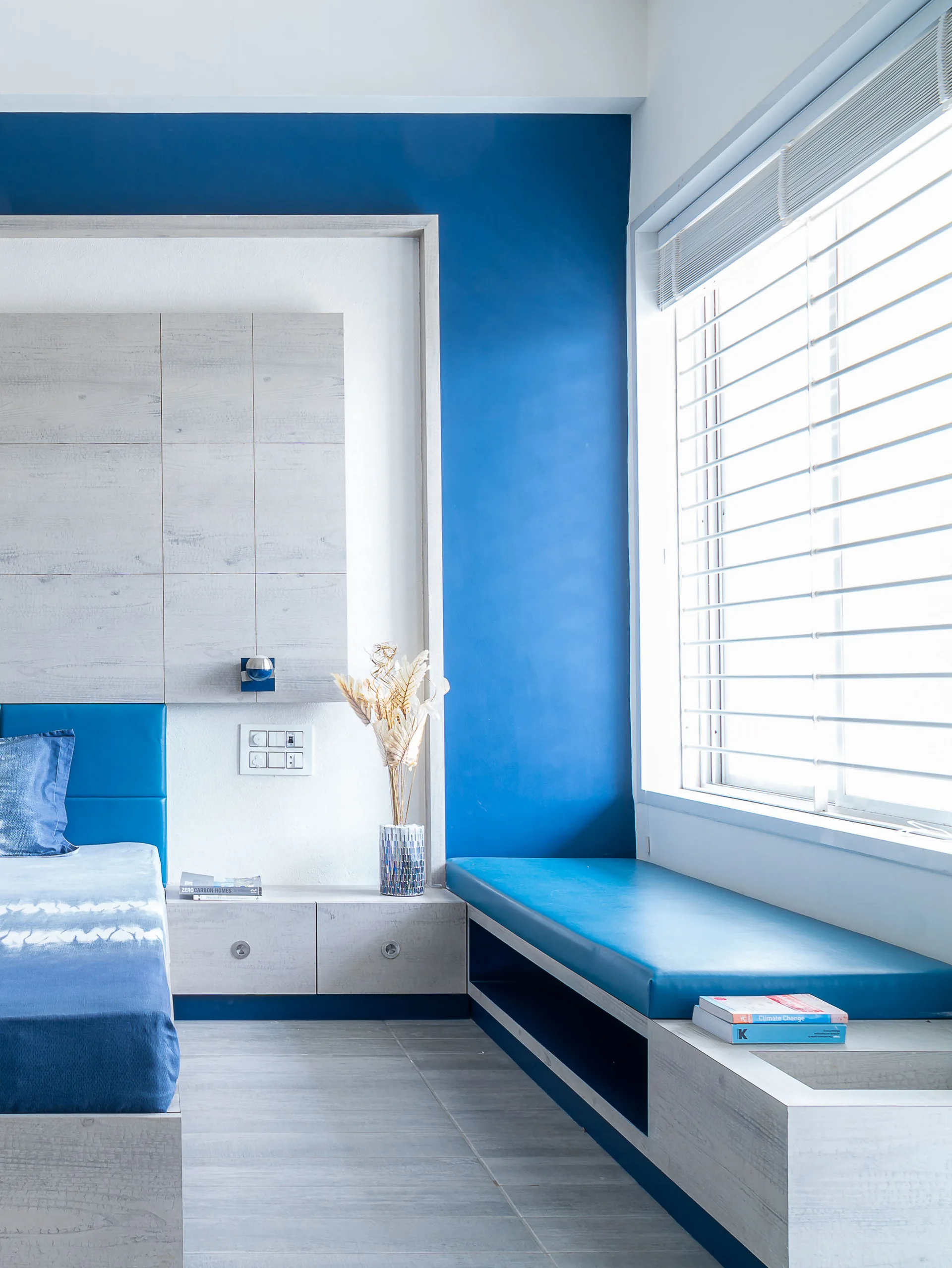 A modern bedroom features a bright blue accent wall, a built-in light wood-grain window seat with a blue cushion, and matching wall panels.