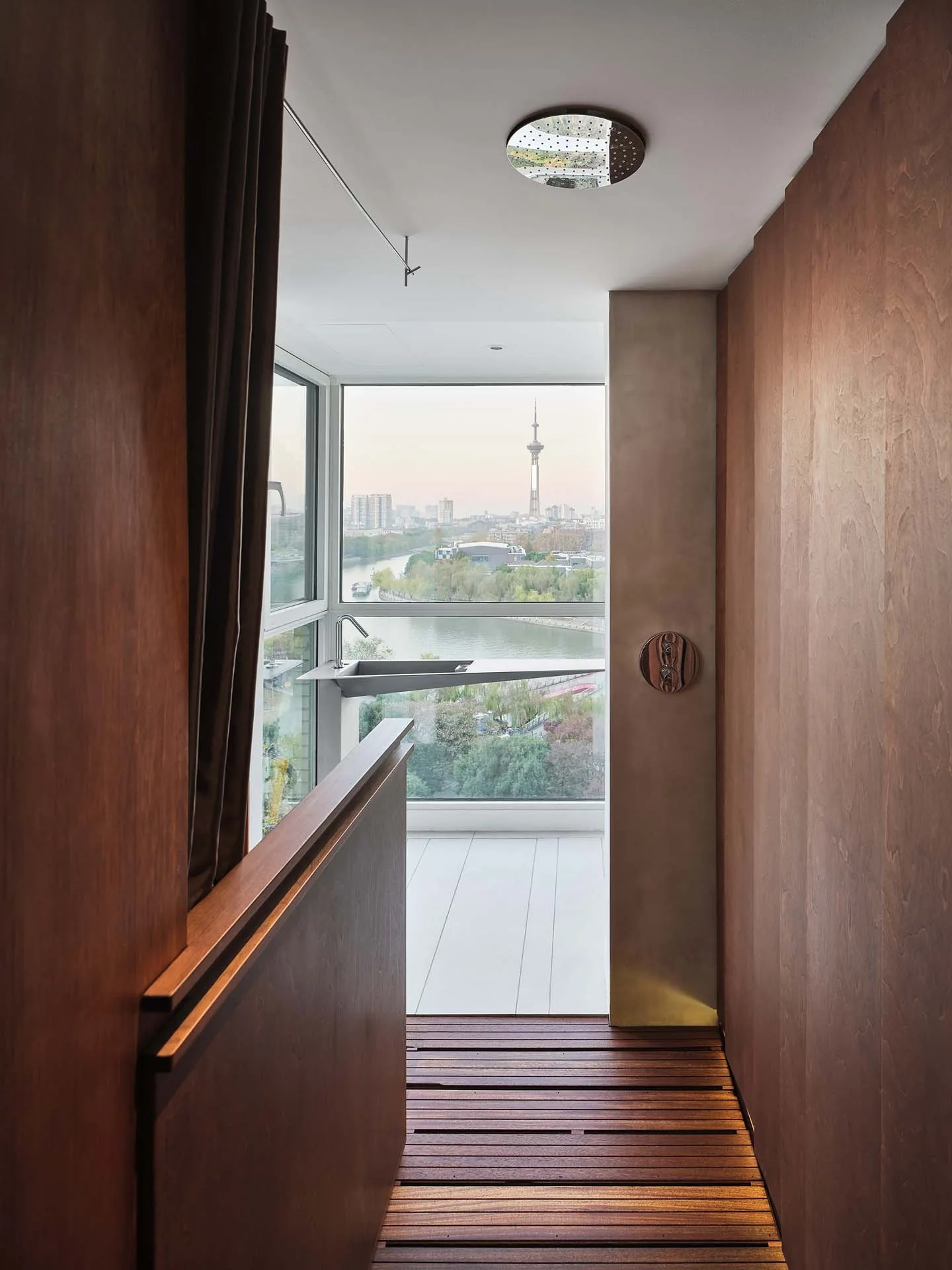 A minimalist shower area featuring vertical wood wall panels, a slatted floor, and a triangular sink overlooking a river and city skyline.