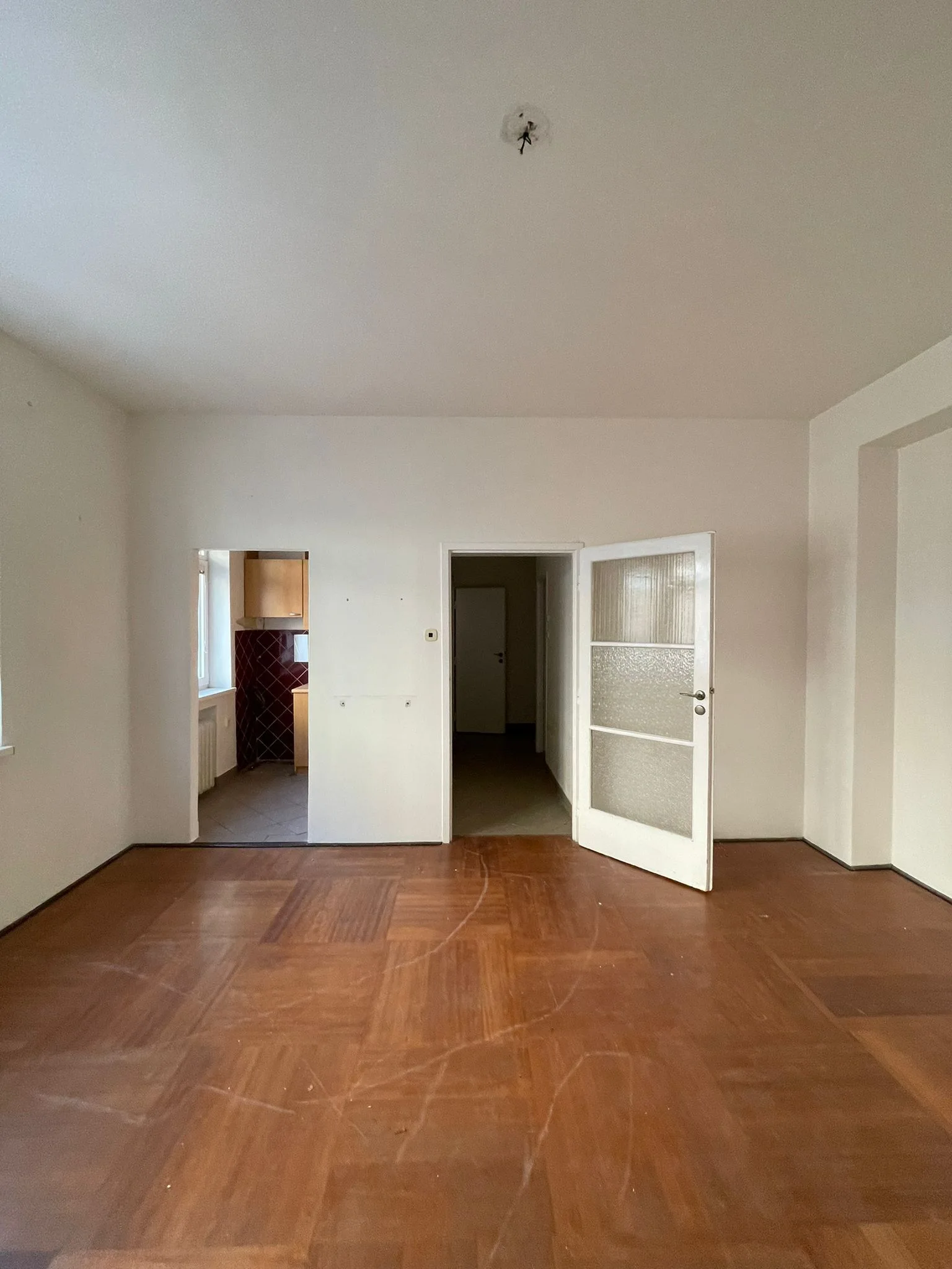 An empty white room with a classic brown parquet wood floor features two doorways, including one with an open white glass-paneled door.