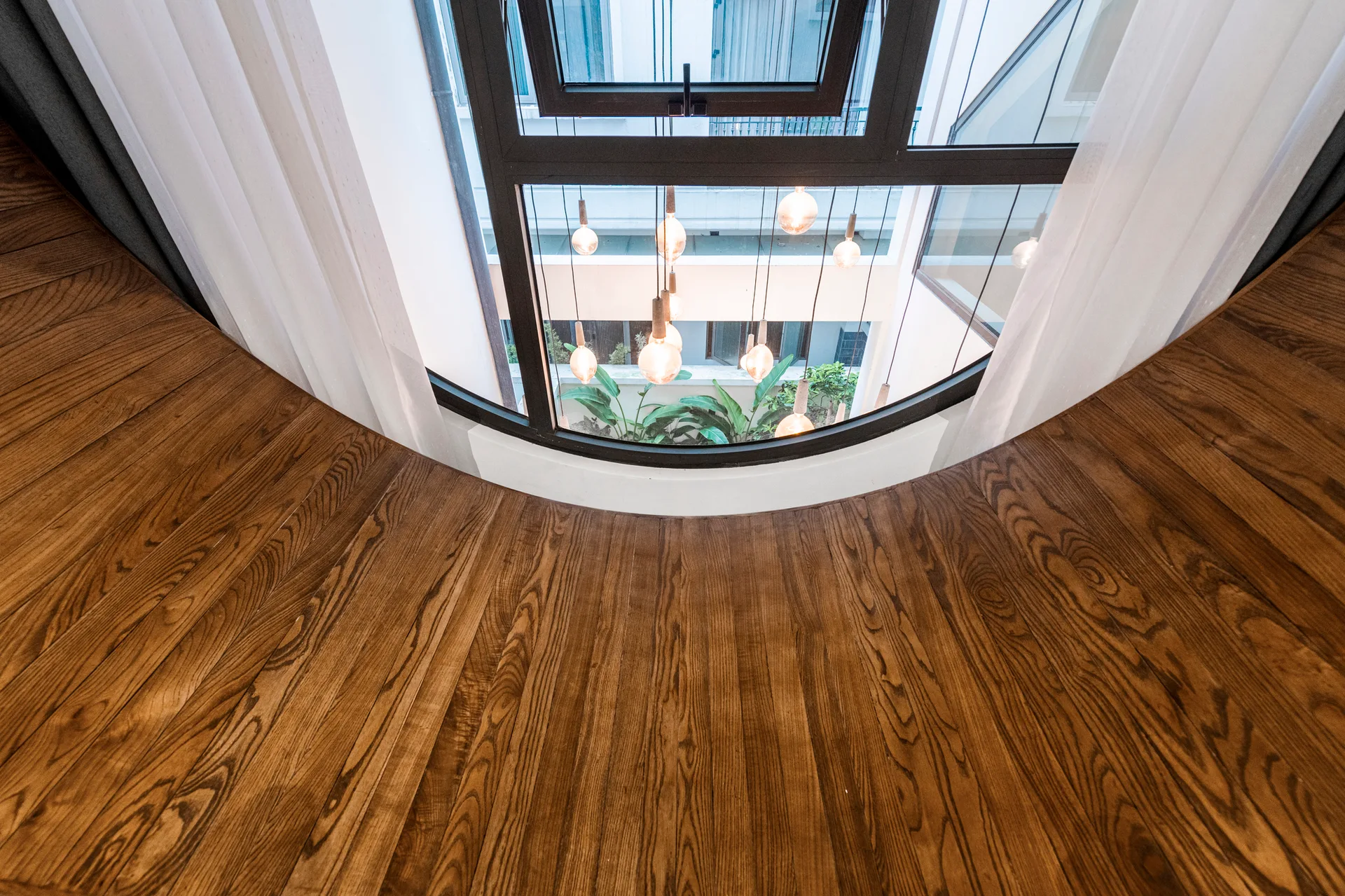 A curved hardwood balcony floor sits before a large window overlooking an indoor courtyard with hanging bulb pendant lights and green plants.