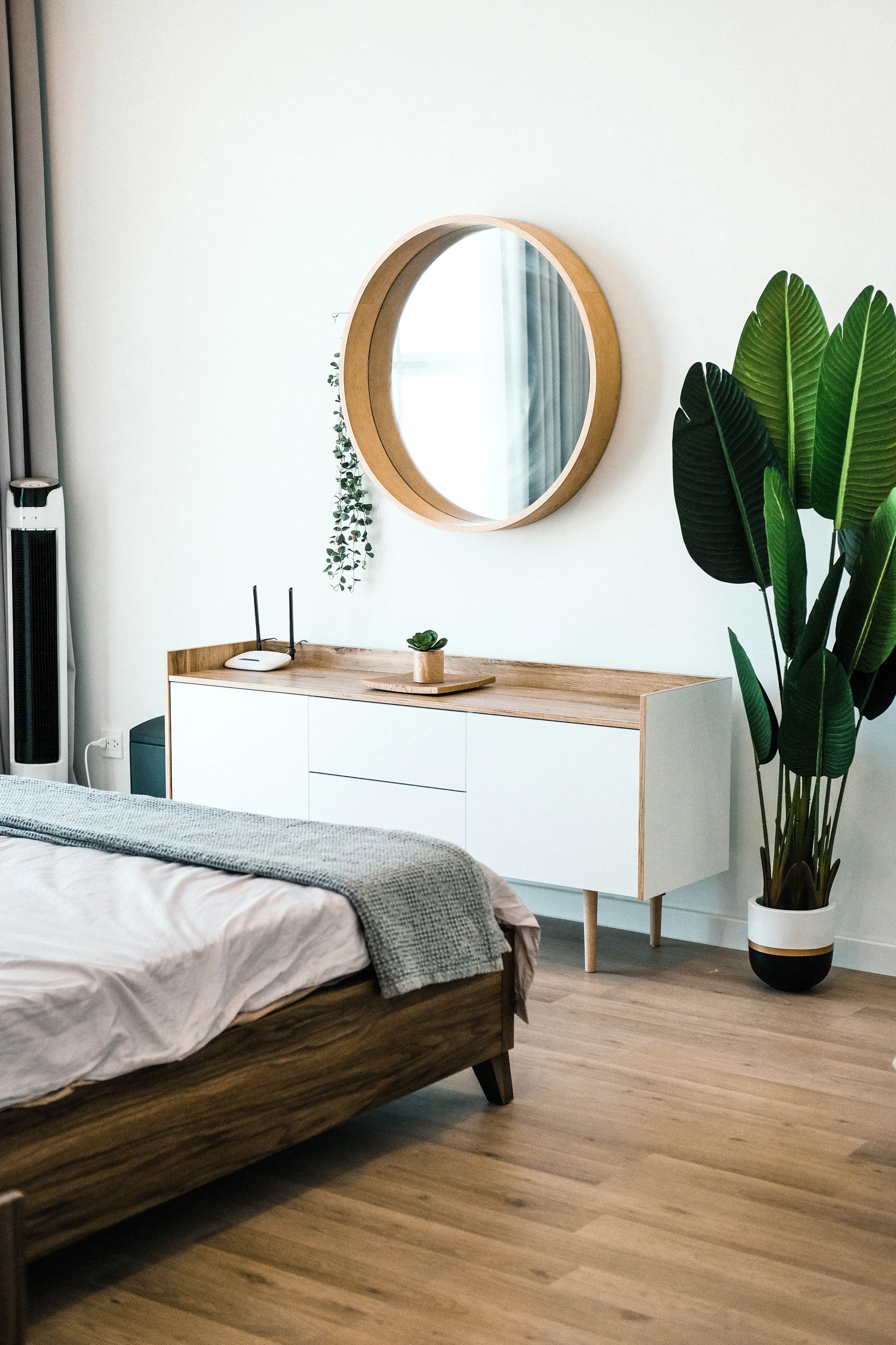 A minimalist bedroom features a wooden bed, a white and wood console table, and a large circular mirror on the wall.