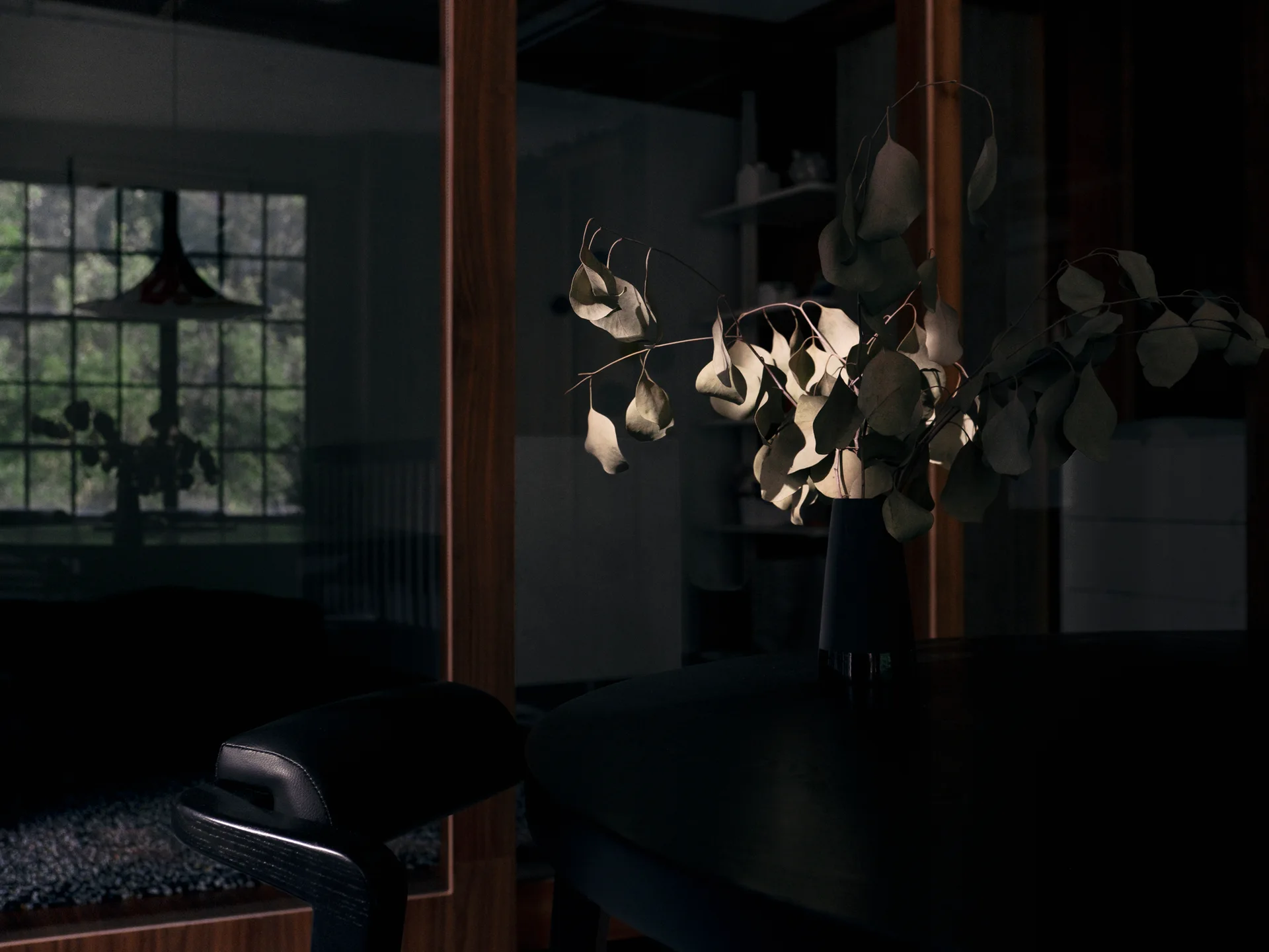 Dried branches in a black vase sit on a dark round table under focused light, backed by a wood-trimmed glass divider reflecting a window.
