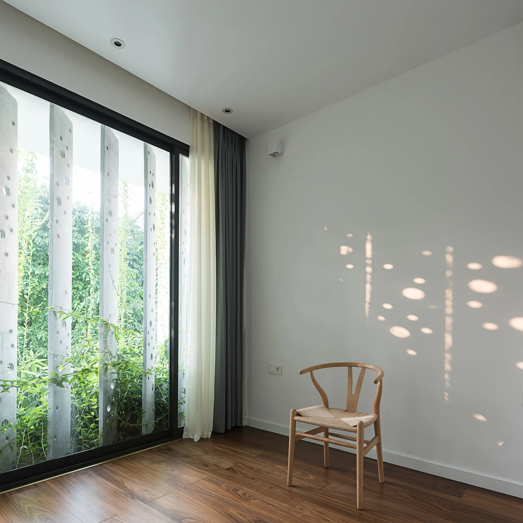 A minimalist room with a light wood wishbone chair on dark wood floors beside a glass door that casts dappled sun shadows on a white wall.