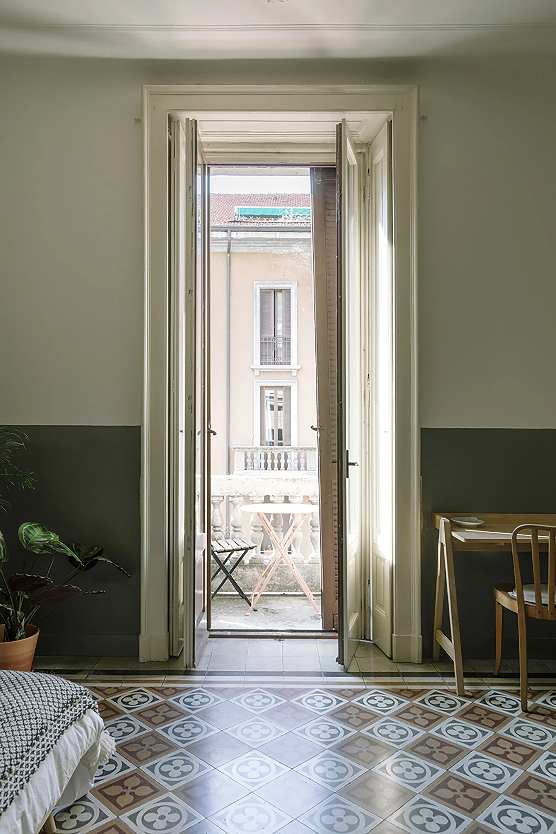 A bedroom features patterned floor tiles, a two-tone white and grey wall, a wooden desk, and open French doors leading to a balcony.
