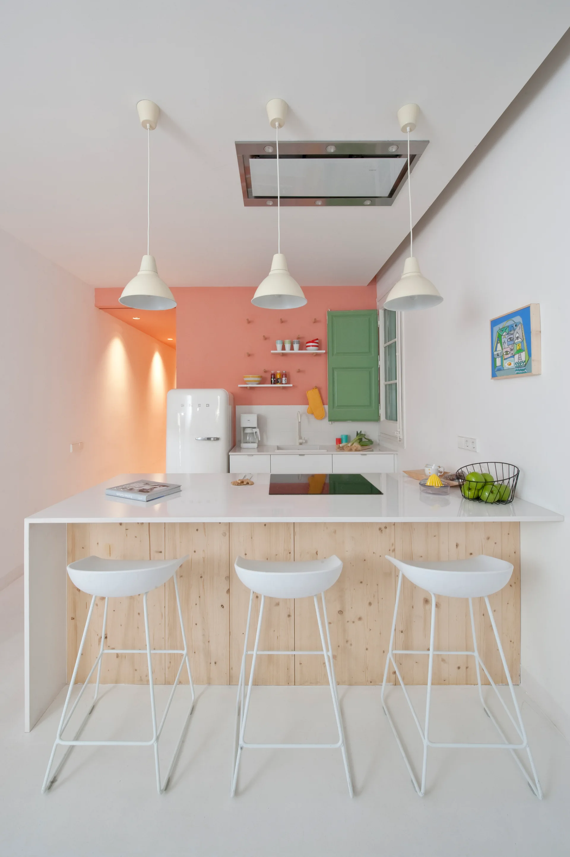 A modern kitchen featuring a white island with a light wood base, three white bar stools, and a pastel coral accent wall.