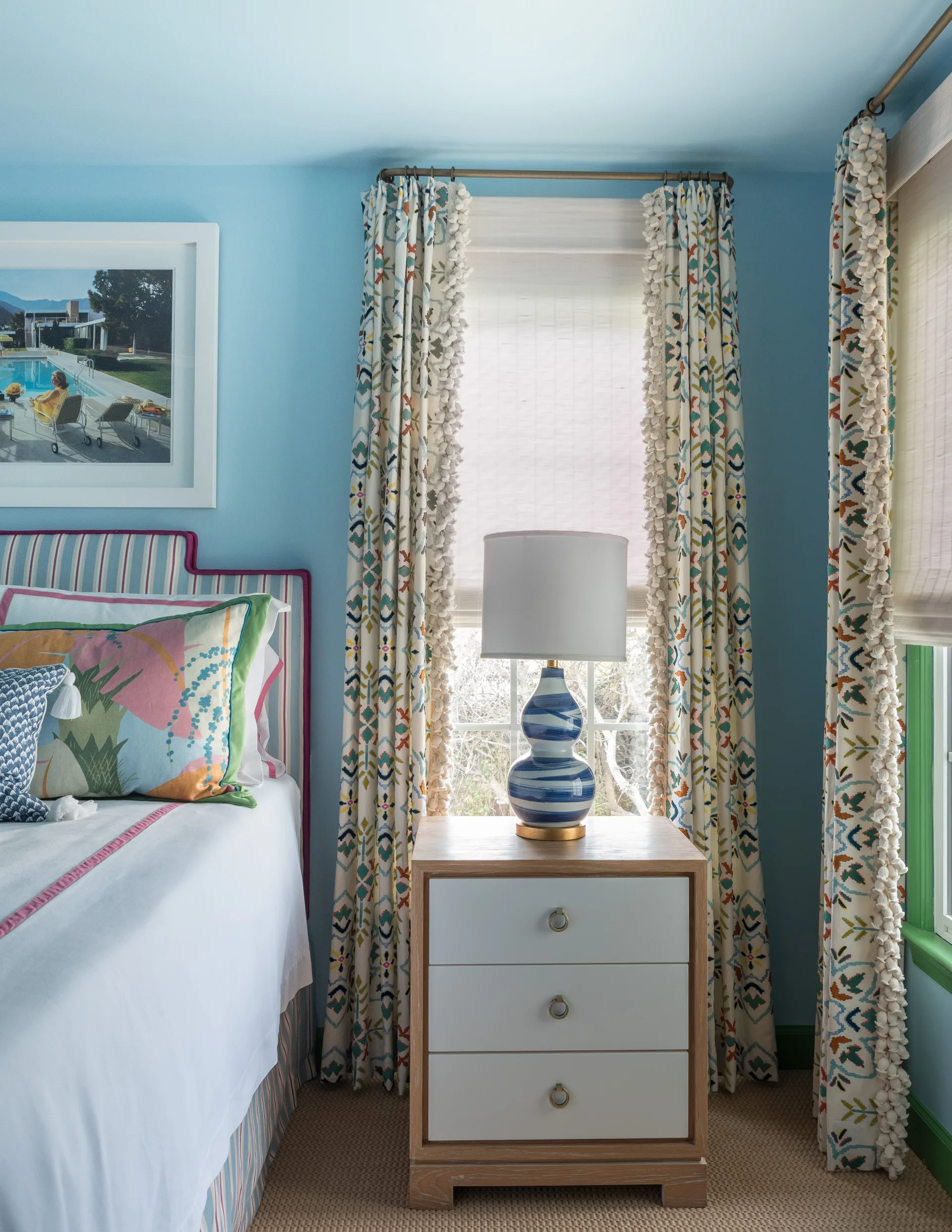 A bedroom corner features a blue and white striped headboard next to a light wood nightstand and window curtains with pom-pom trim.